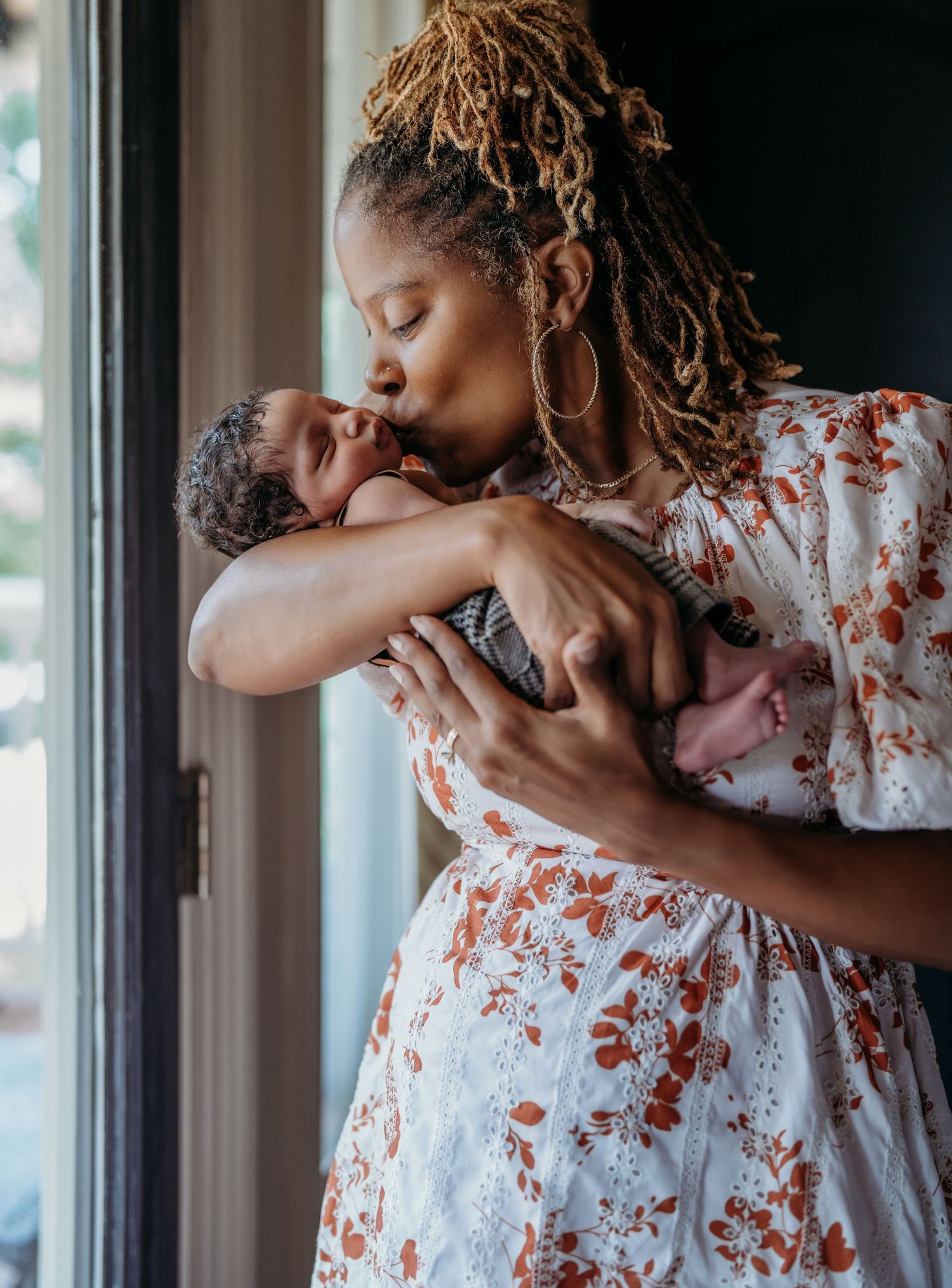 A tender moment by a window as a mother in a floral dress cradles and kisses her newborn baby.
