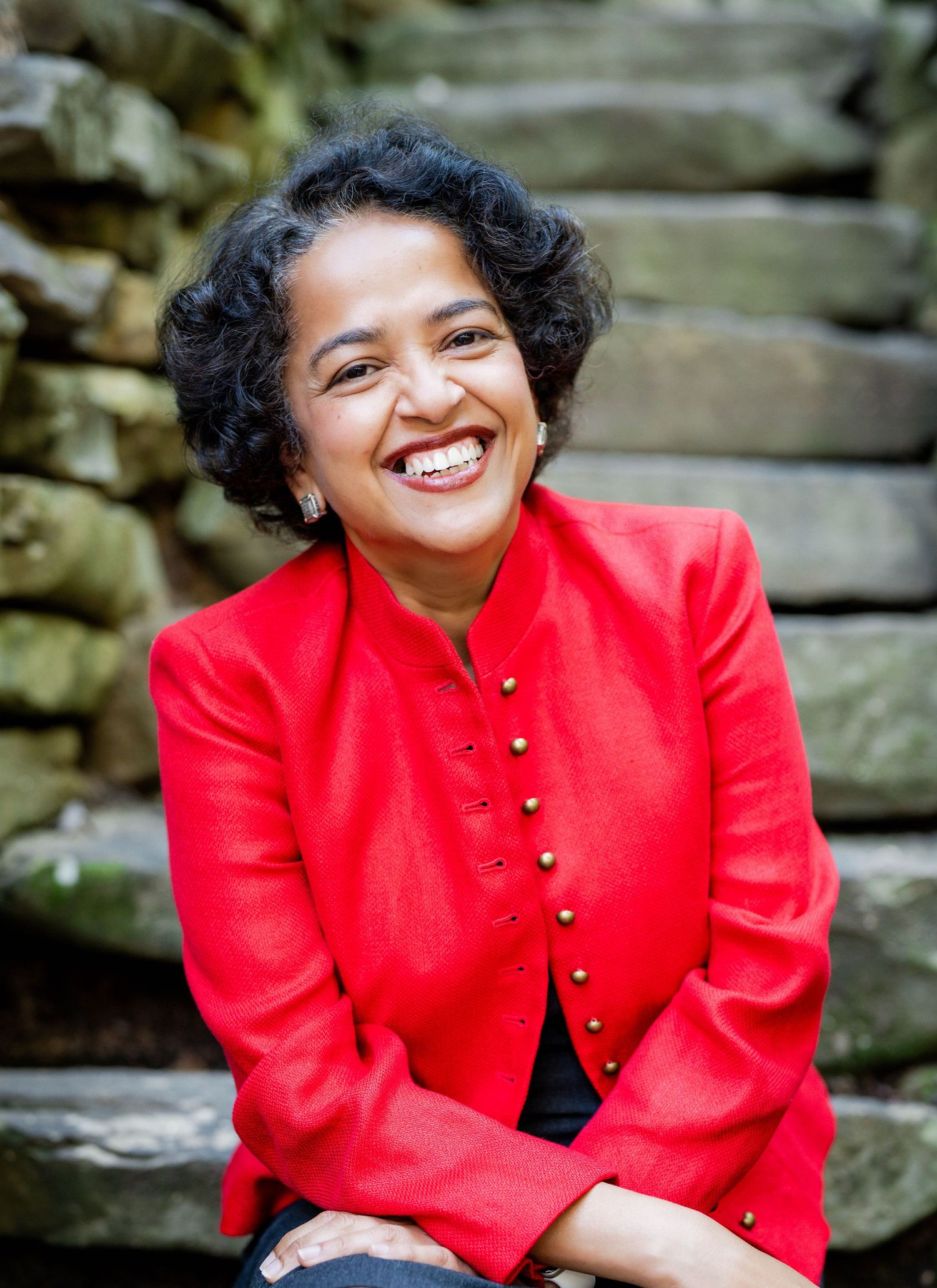 Person in vibrant red buttoned jacket smiles warmly while seated against stone wall background.