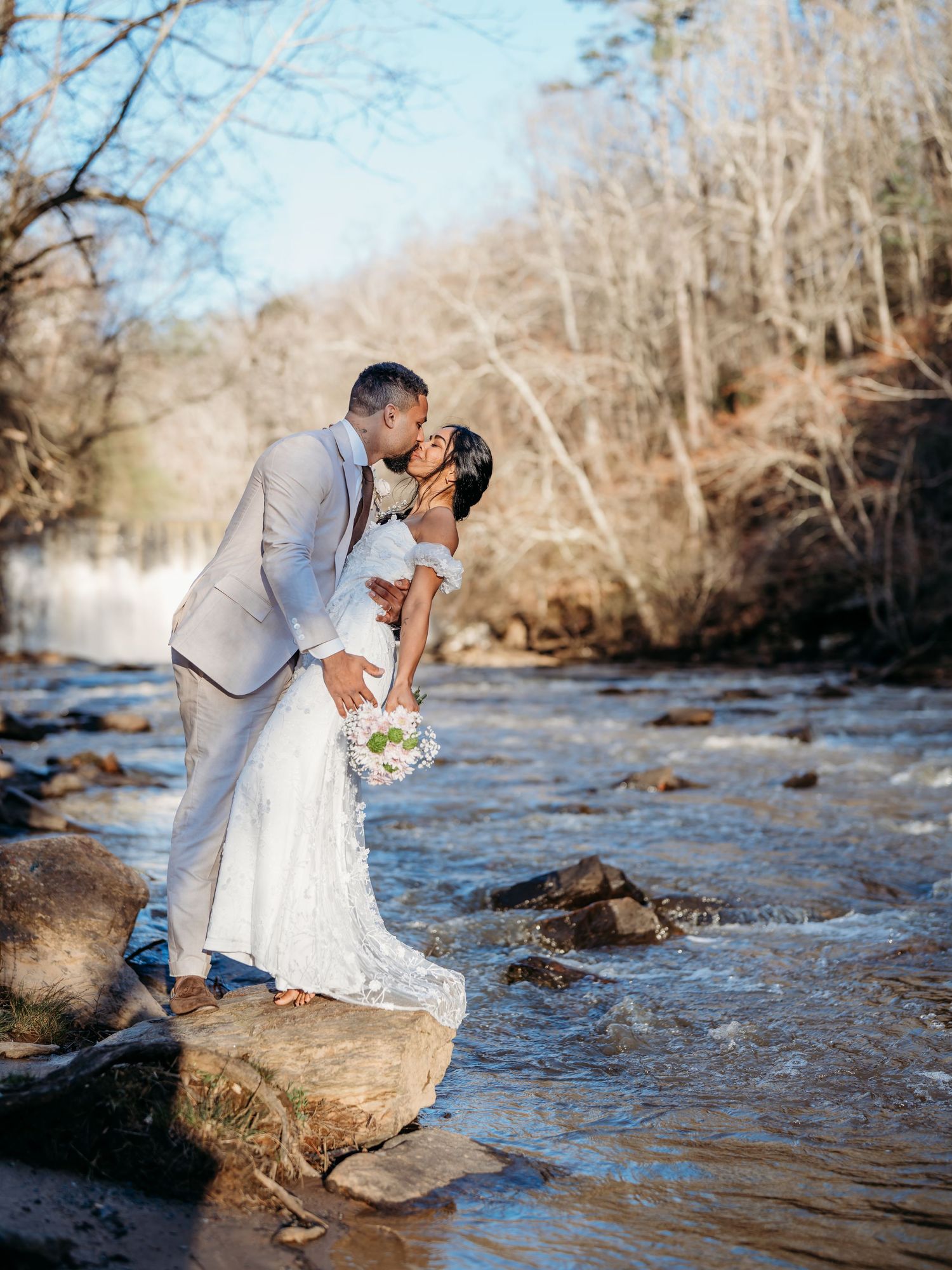 A romantic wedding couple embraces on rocks beside a flowing river during an outdoor photo session.