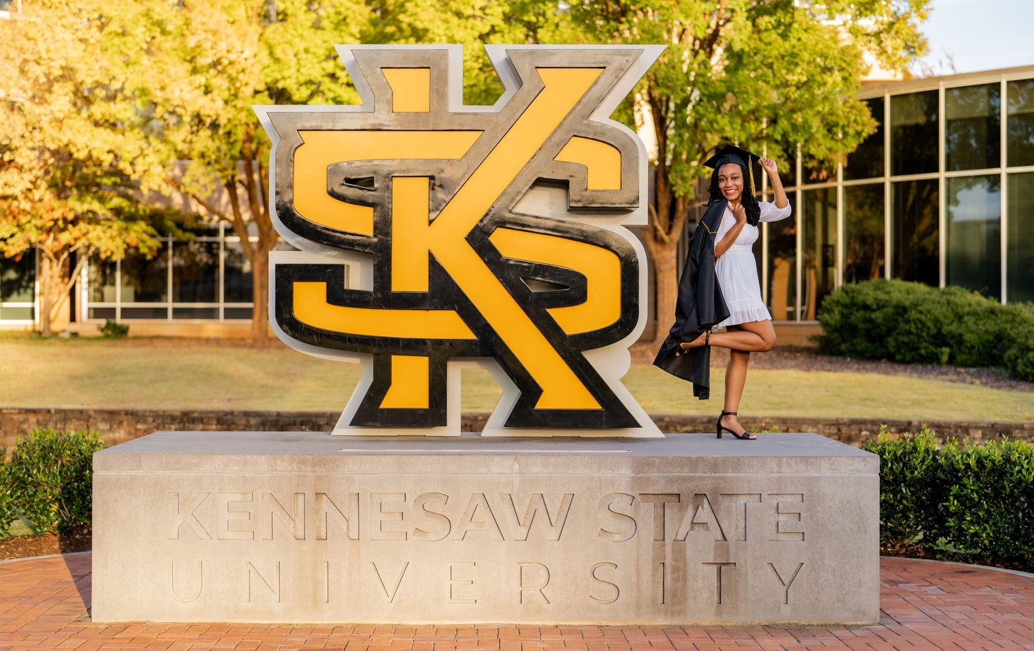 A student poses by the large gold logo sign at Kennesaw State University during autumn with yellow trees in background.