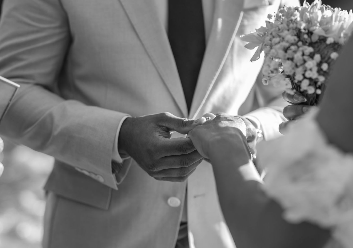 Black and white photo showing hands exchanging rings during a wedding ceremony with flowers in view.