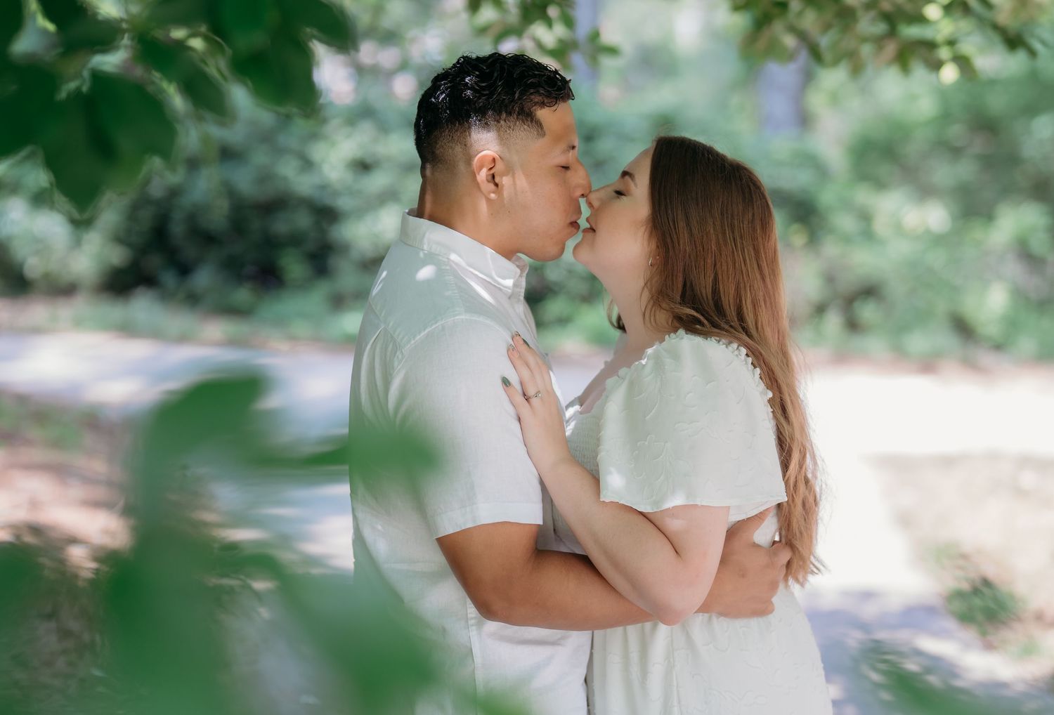 Romantic couple in light summer outfits sharing tender kisses during an outdoor photoshoot in a sunlit wooded setting.