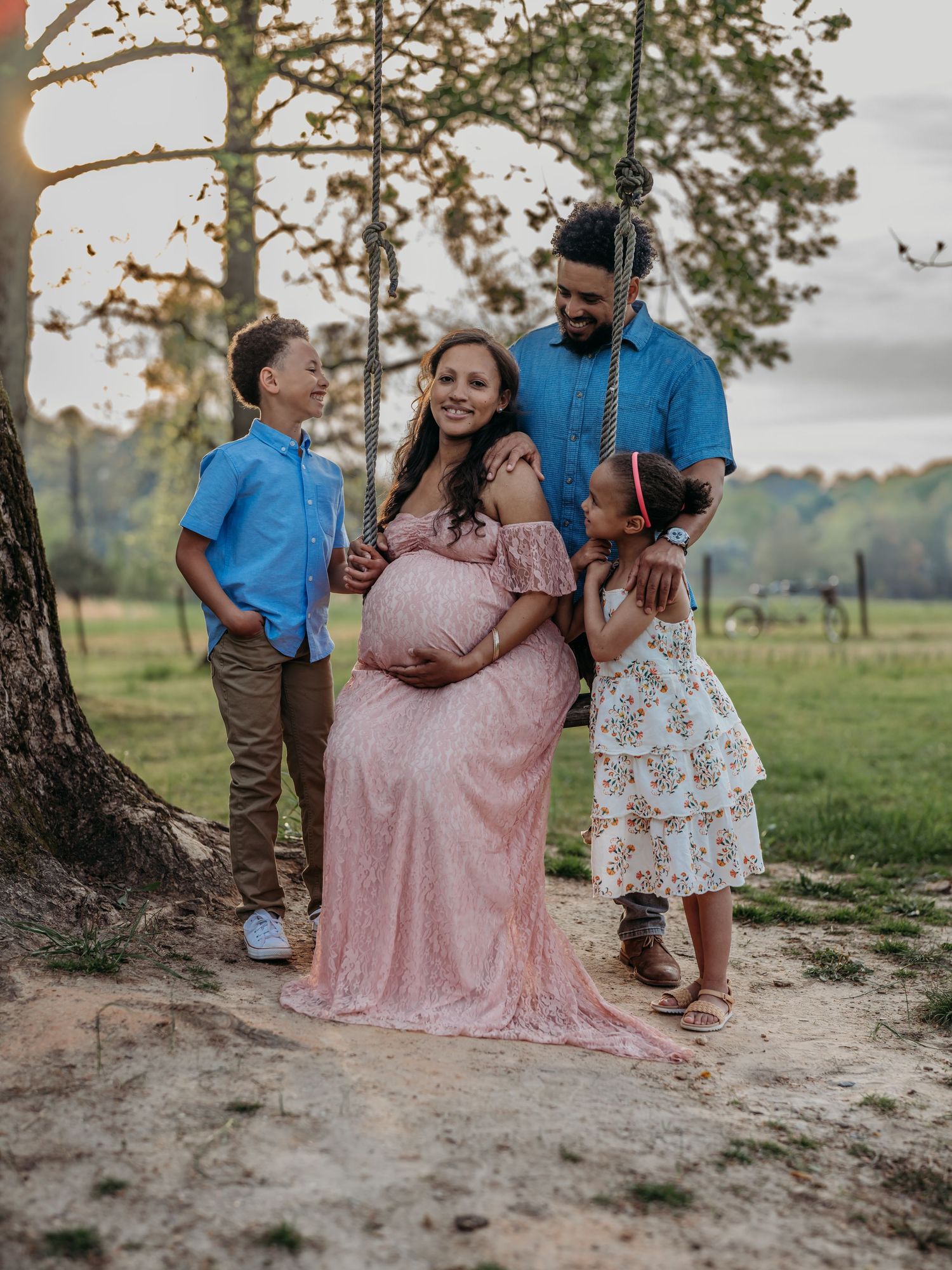 A family poses together under a tree in an outdoor setting with a pregnant woman in a pink dress.