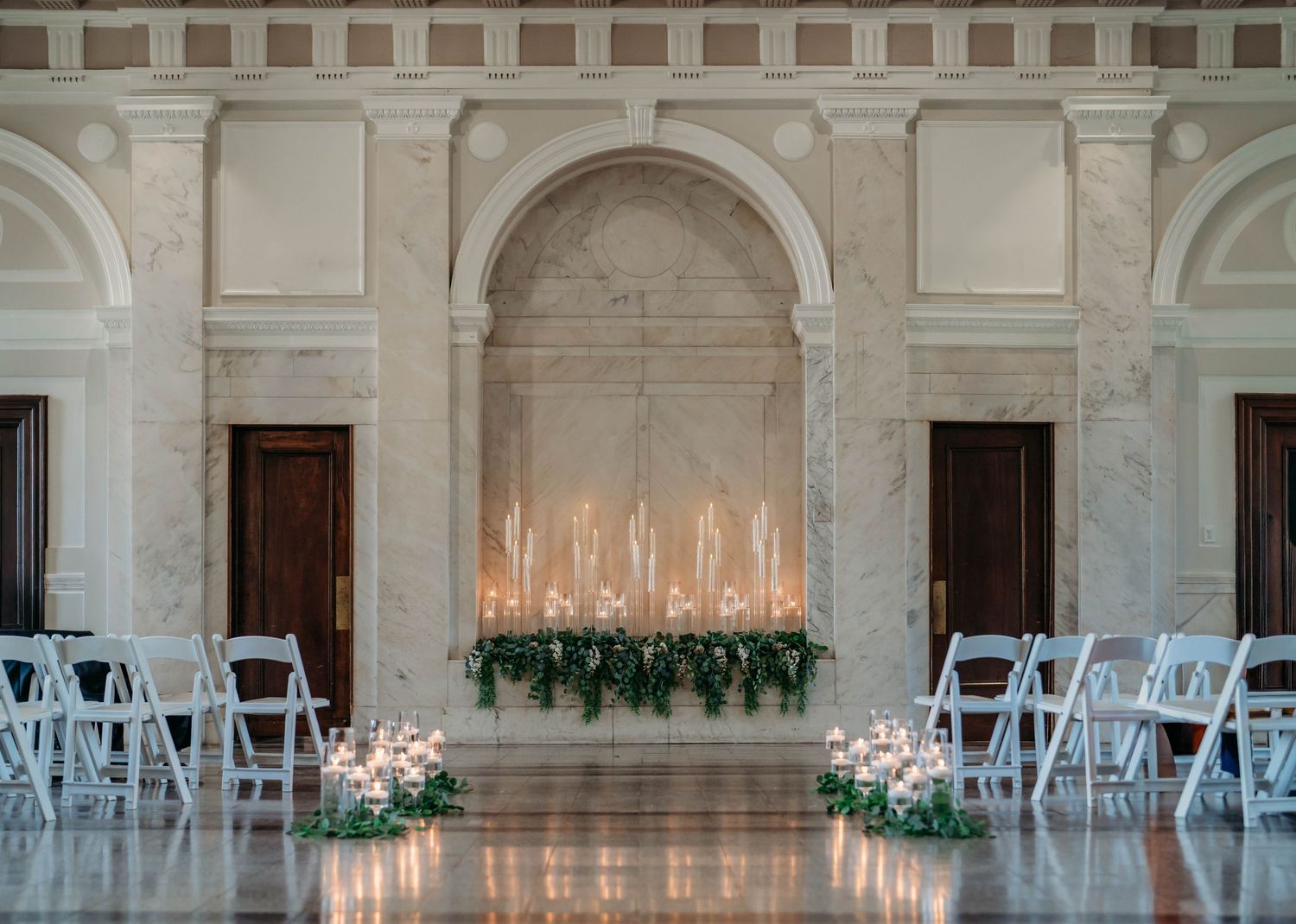 White folding chairs line a grand marble hallway with ornate columns and candelabras for an elegant indoor ceremony setup.