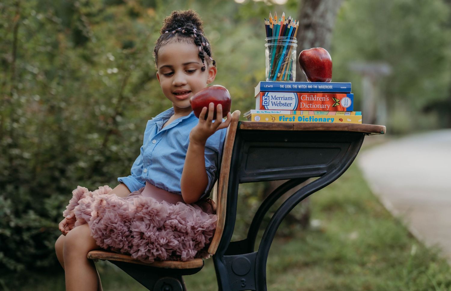 Young girl in tutu sits at vintage school desk outdoors, holding apple beside stacked books and pencils.