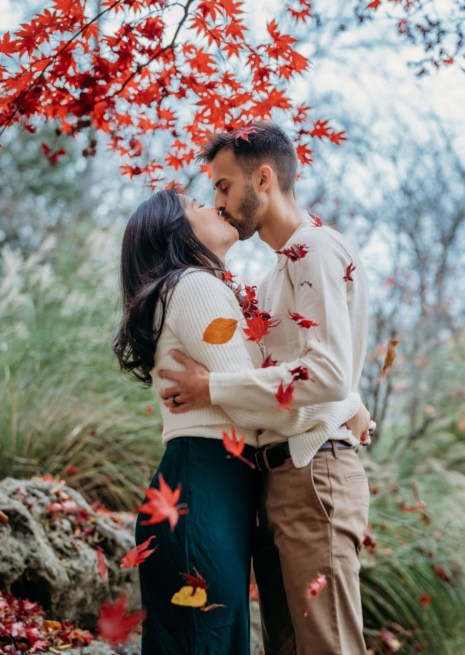 A romantic couple sharing a tender moment under vibrant red autumn maple leaves in a garden setting.