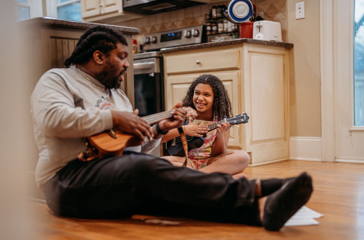 Two people sit on hardwood floor in kitchen playing acoustic instruments together while smiling and enjoying music.