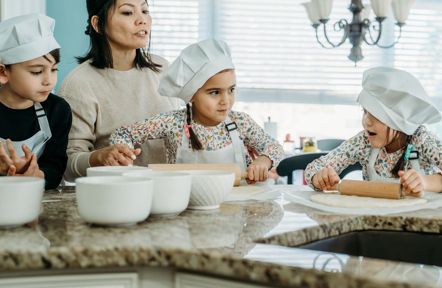 Children in chef hats learn to bake at a granite kitchen counter with mixing bowls and materials ready.