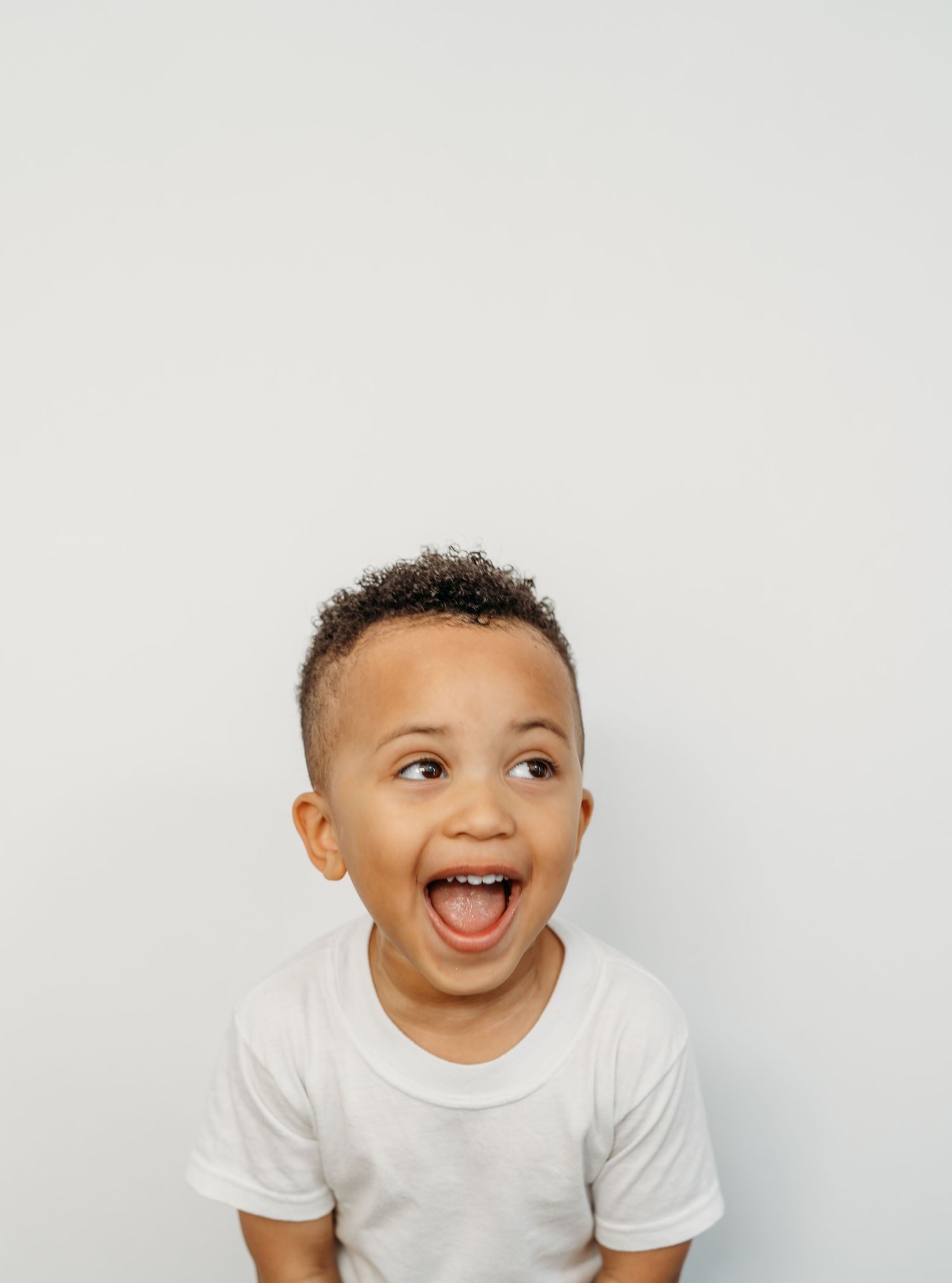 A happy baby in a white t-shirt smiles and laughs while playing against a light gray background.