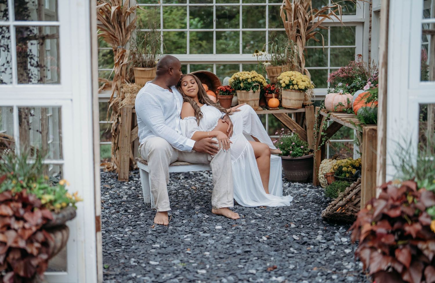 A couple embraces in a romantic outdoor garden setting with white greenhouse windows and dried plants in the background.