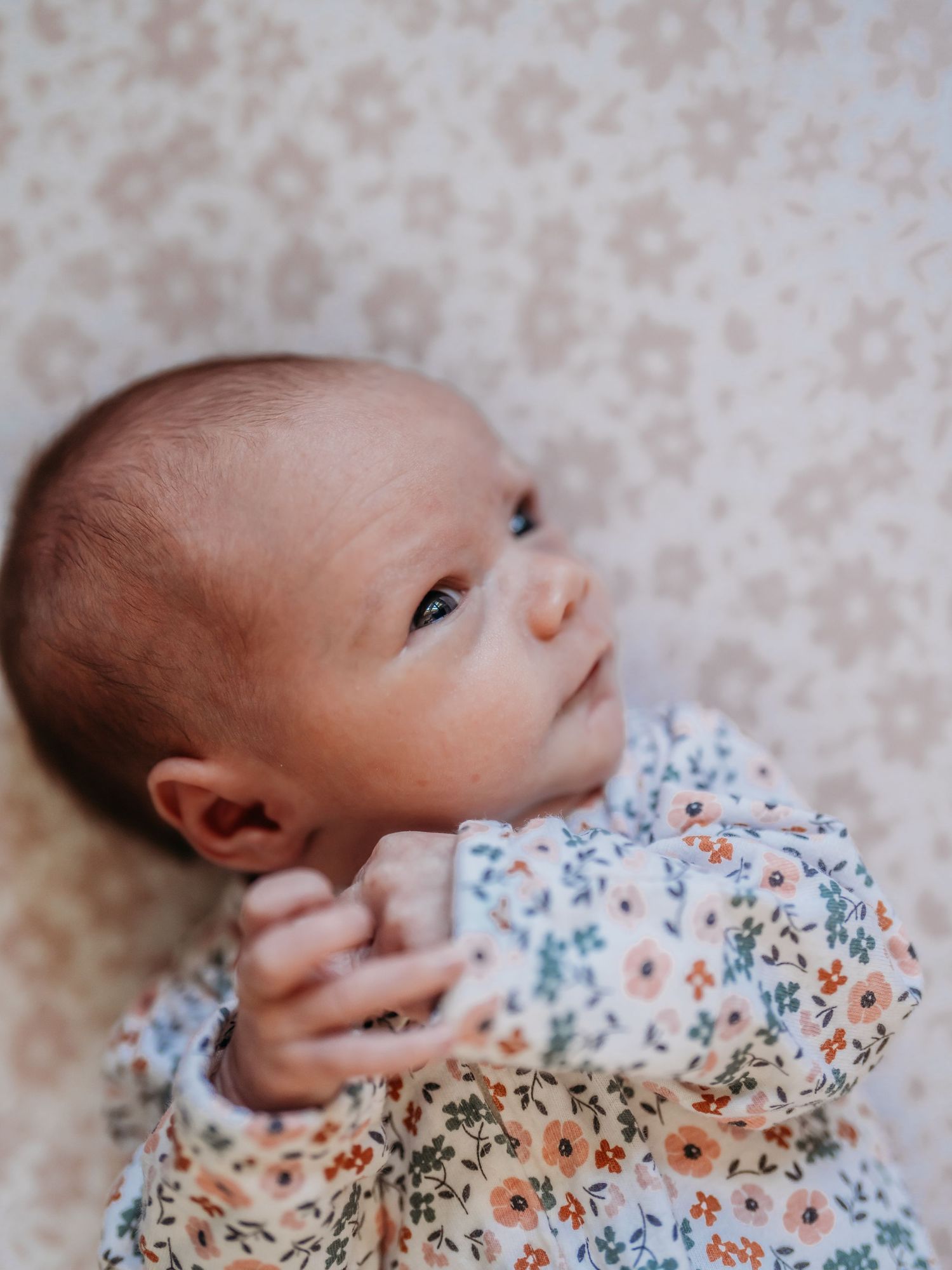 Baby lying down wearing a delicate floral print dress in white, blue and orange tones against a blush background.