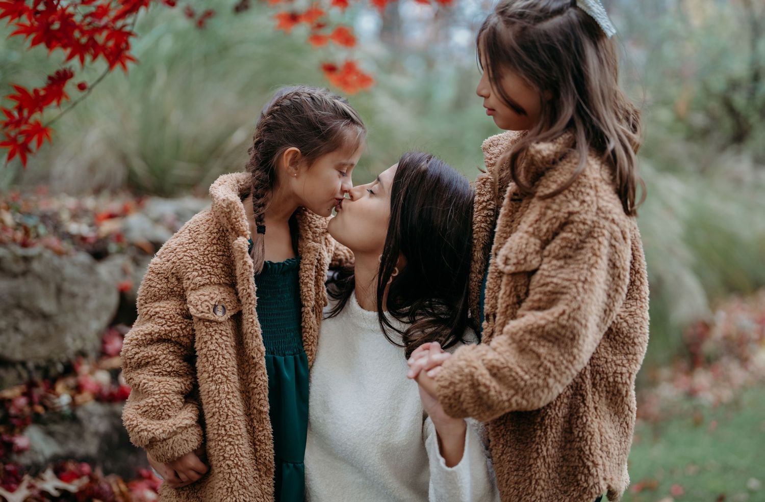 A couple wearing cozy brown teddy coats embrace in a romantic autumn setting with red foliage in the background.
