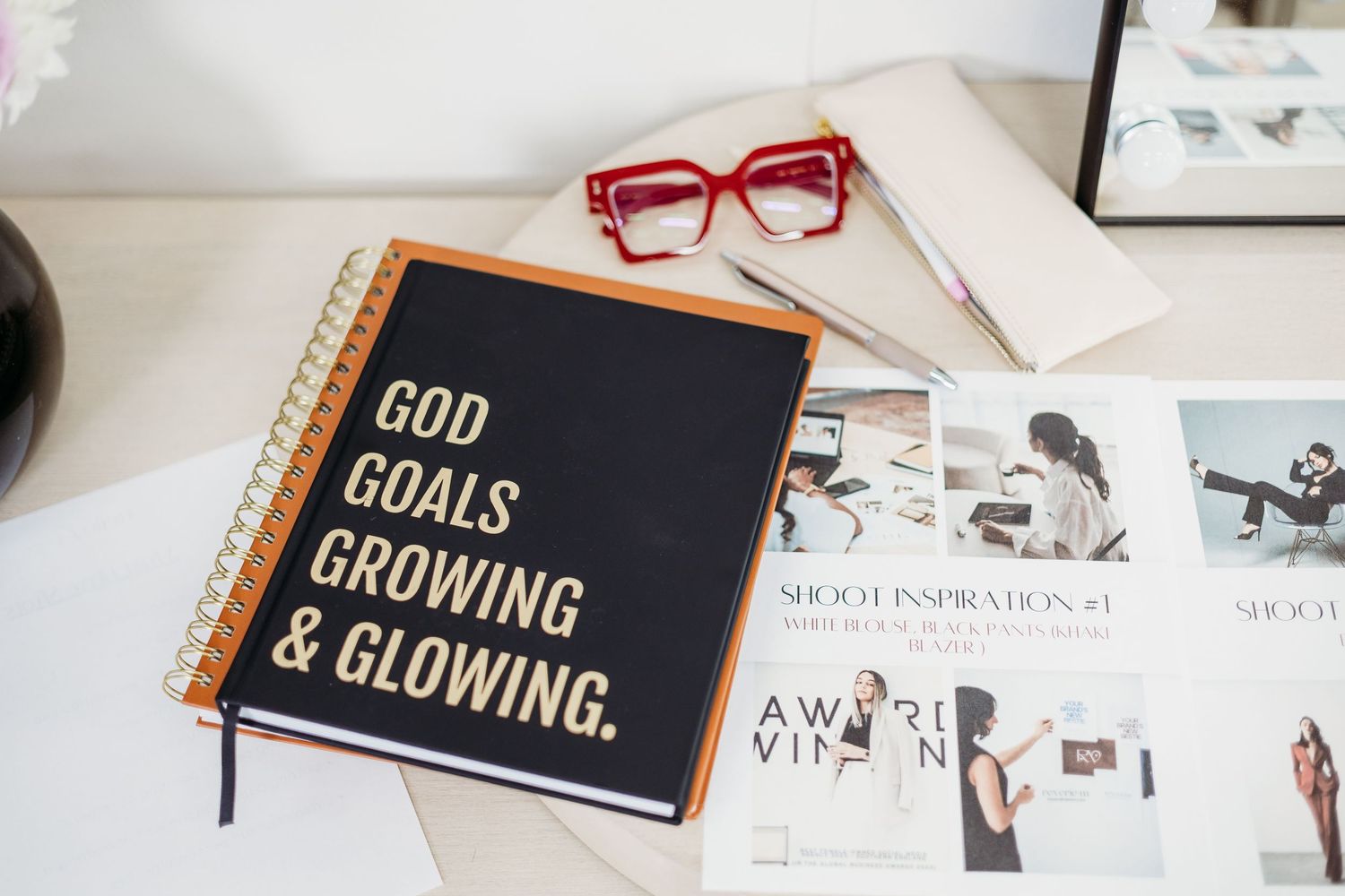 Black motivational notebook with red glasses and photos on white desk displaying 'God Goals Growing & Glowing' text.