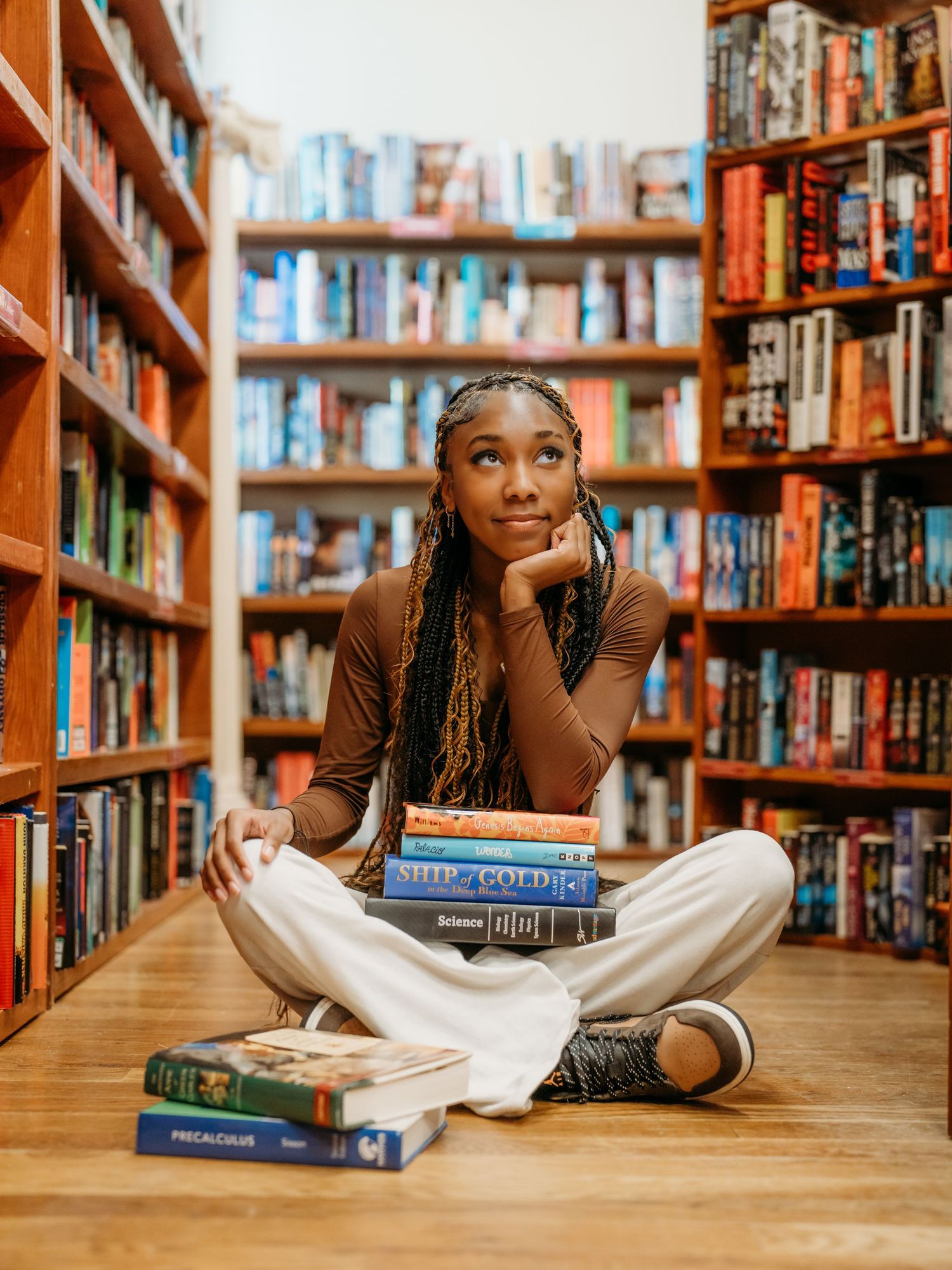 Young reader sitting cross-legged on the floor of a library or bookstore surrounded by bookshelves and stacked books.