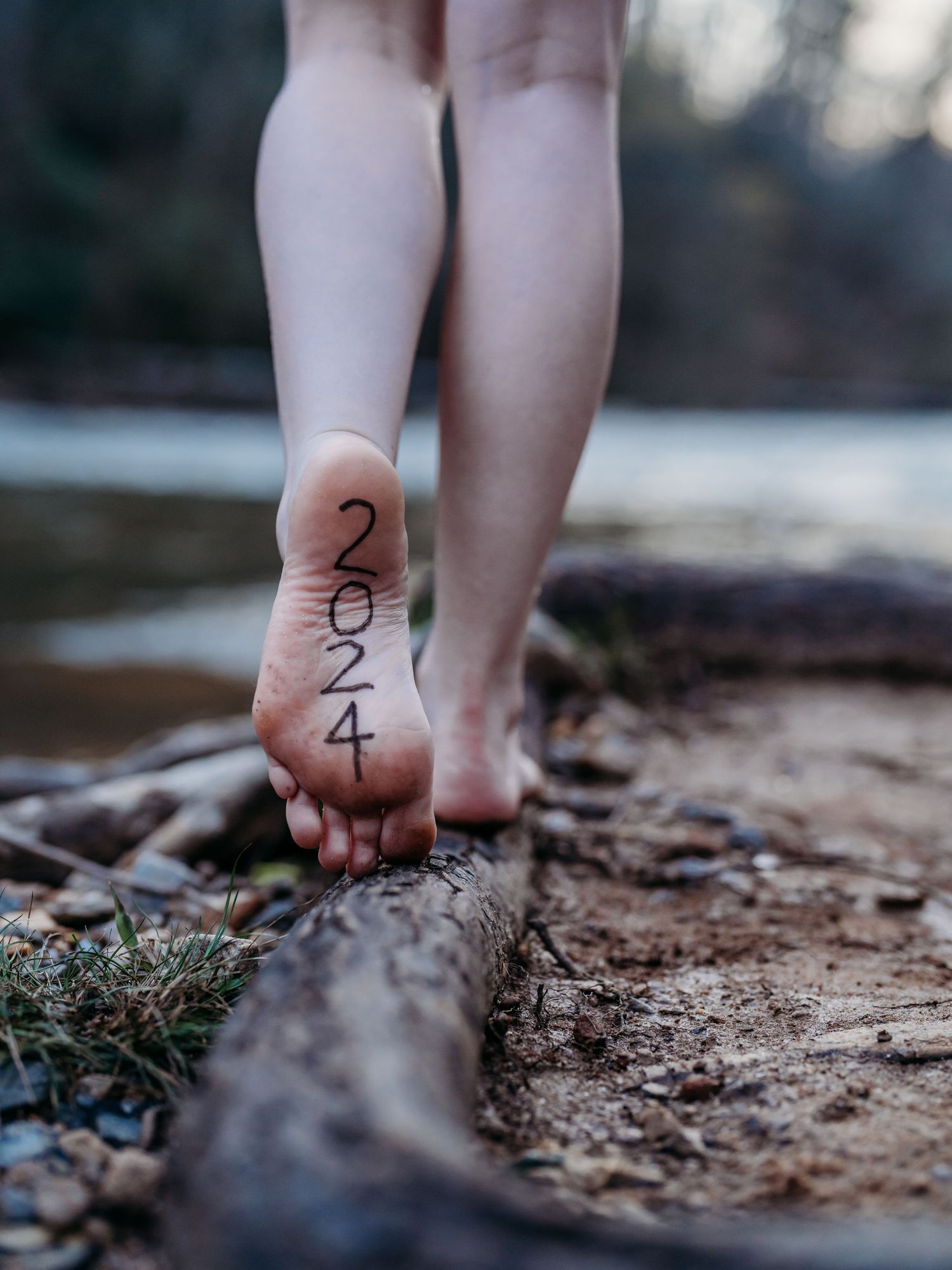 Barefoot person balances on a log with numbers 2-3-4 written on their sole in a natural outdoor setting.