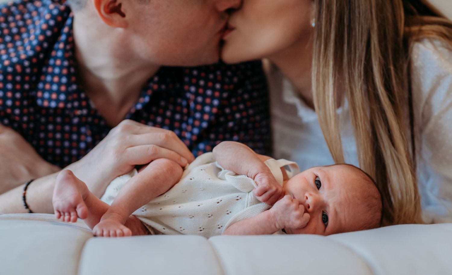 A baby in white clothing lies between two adults sharing an intimate moment on white bedding.