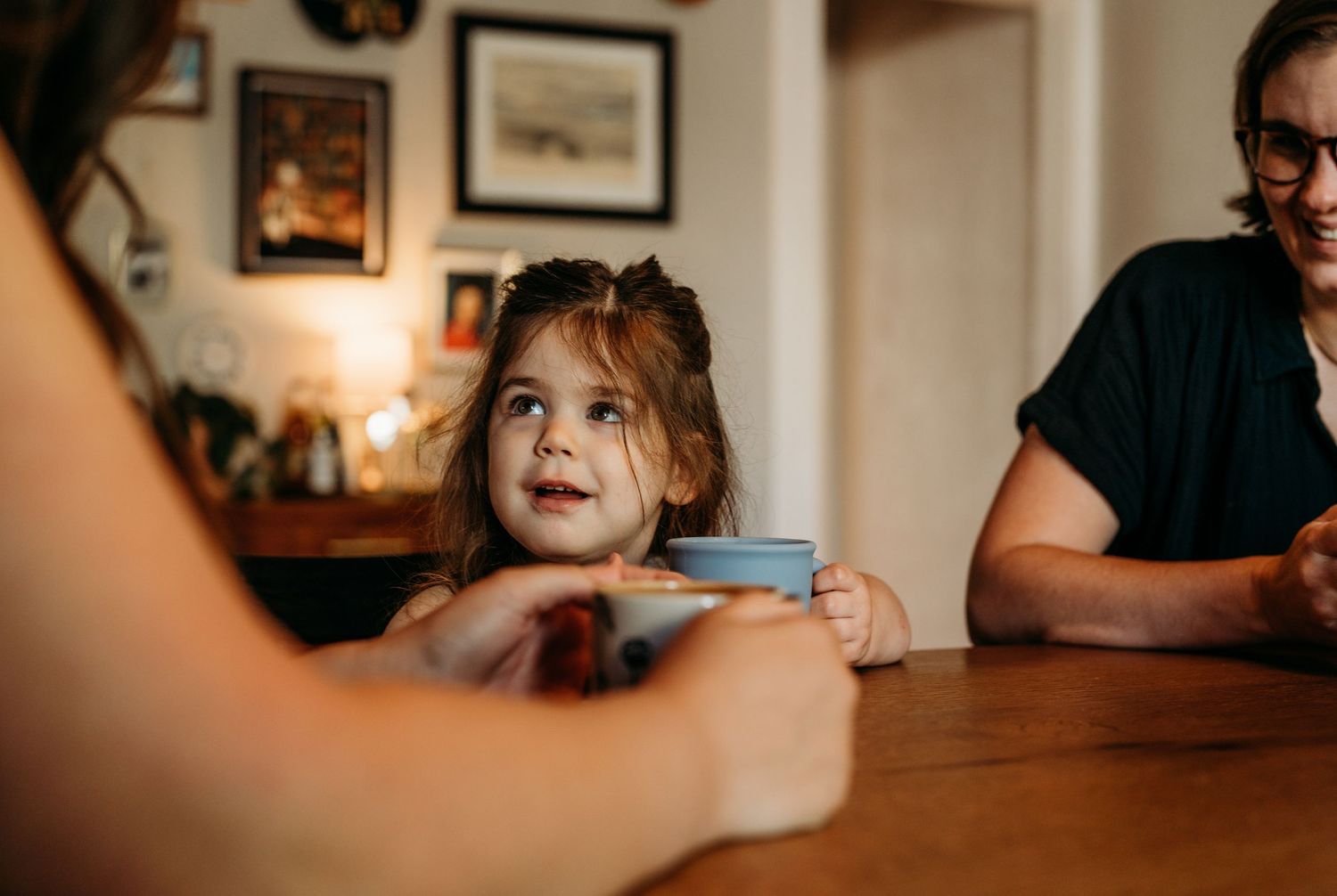 A young child sits with someone at a wooden table during mealtime in a warm, cozy home setting.
