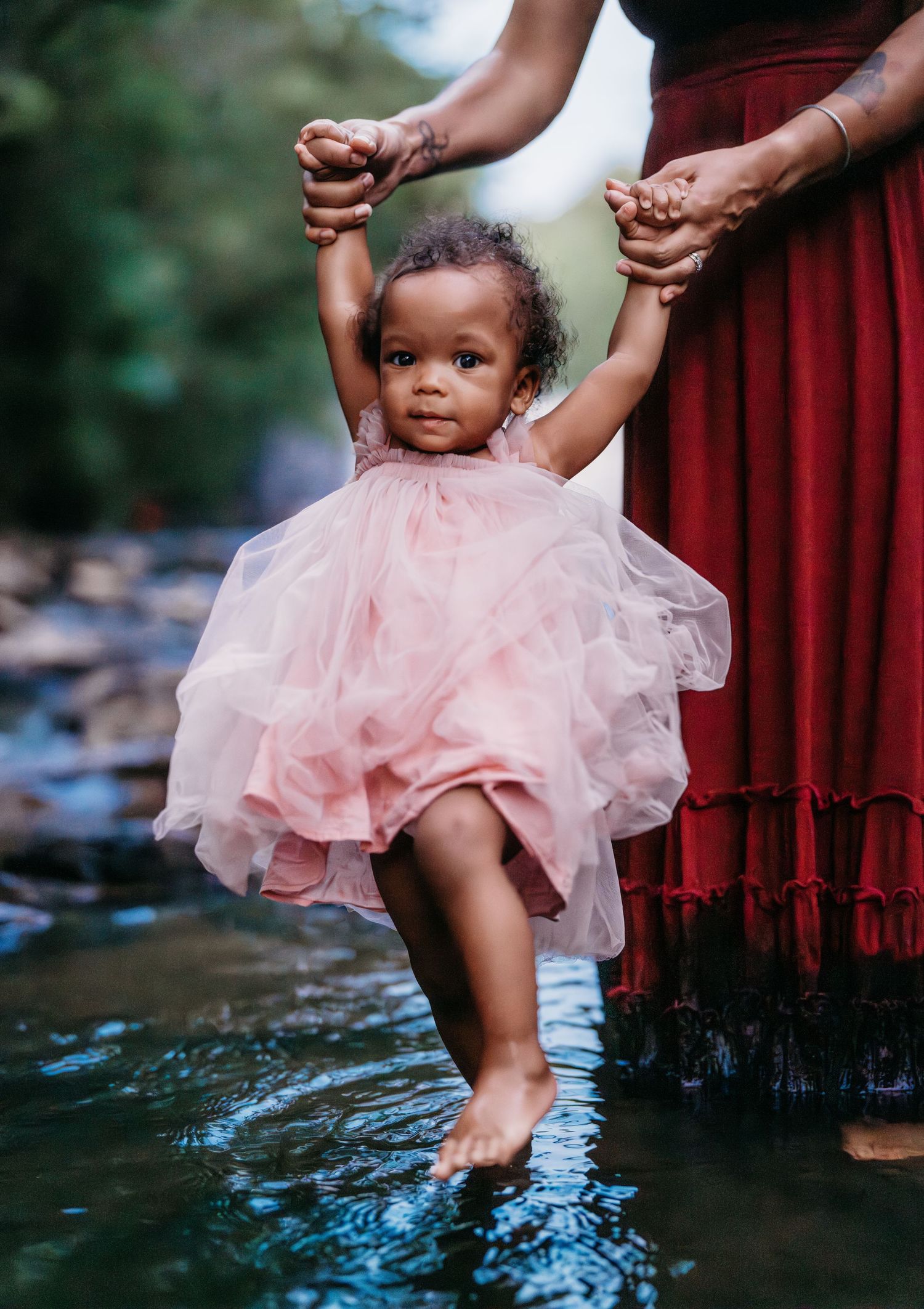 A small child in a pink tulle dress dips their toes in water while being supported by hands in a flowing red dress.