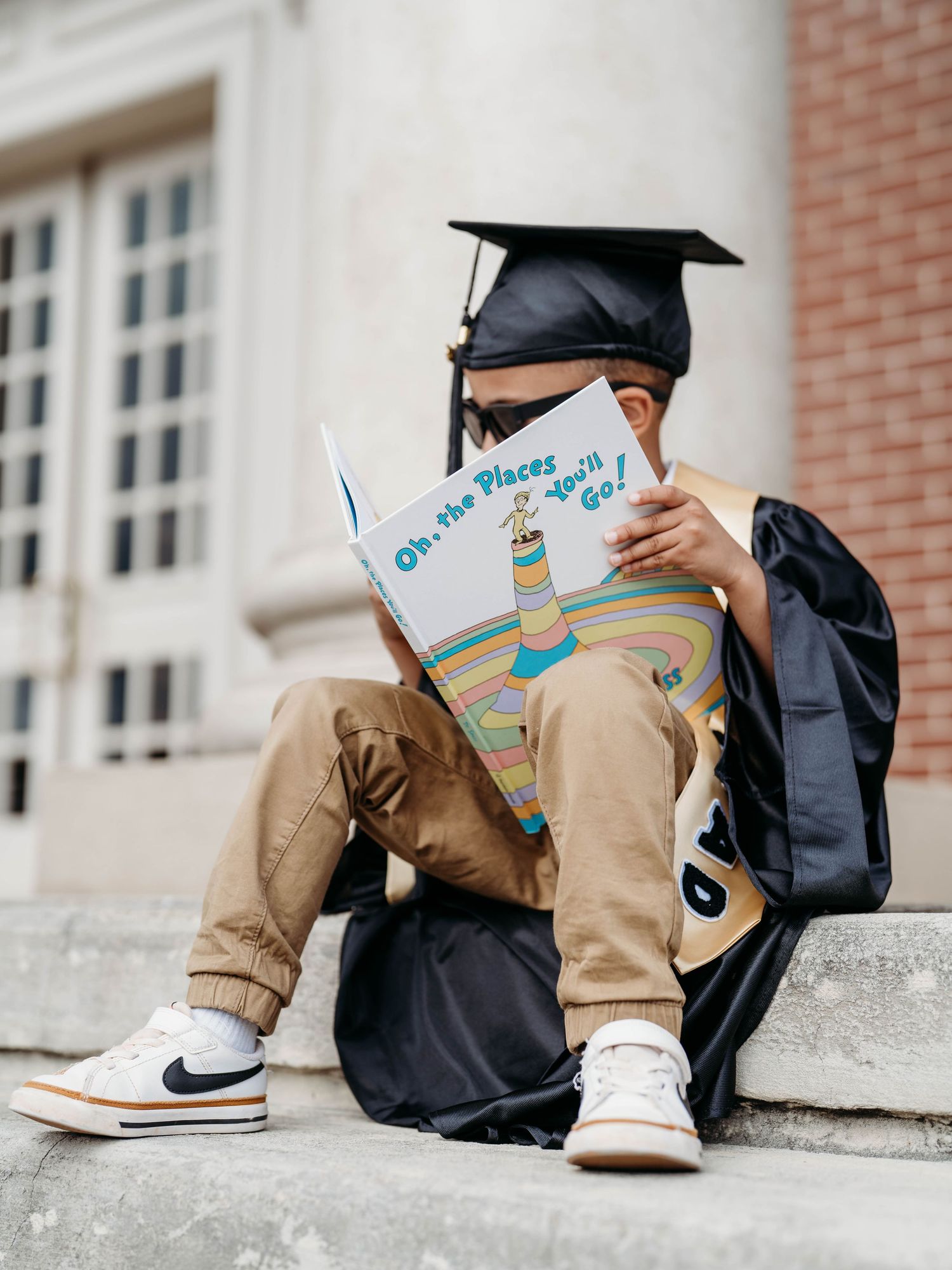 Child in graduation cap reads Oh, the Places You'll Go by Dr. Seuss while sitting on steps with backpack.