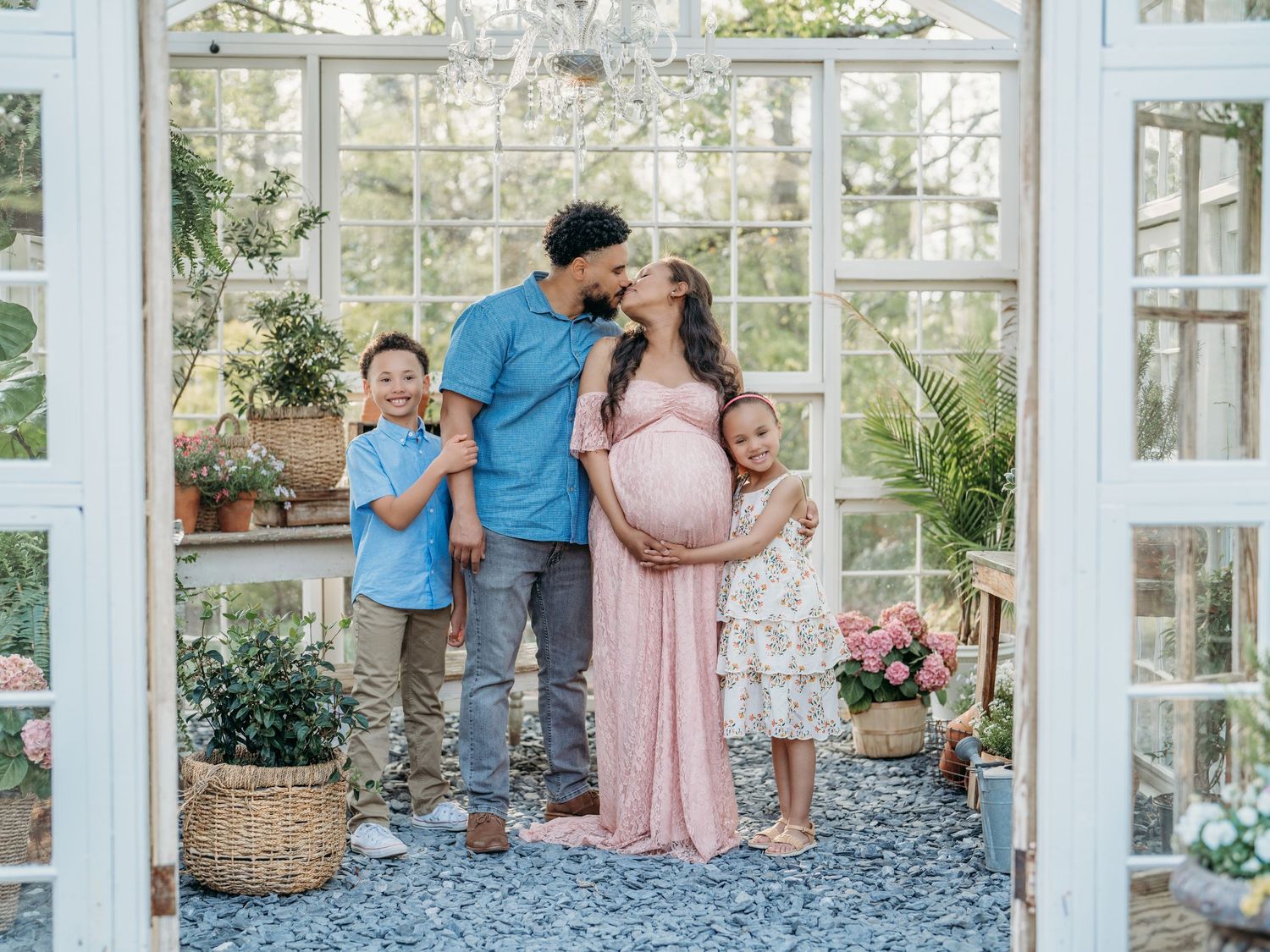 A family in coordinating pink and blue outfits poses together in a bright greenhouse setting with white framed windows.