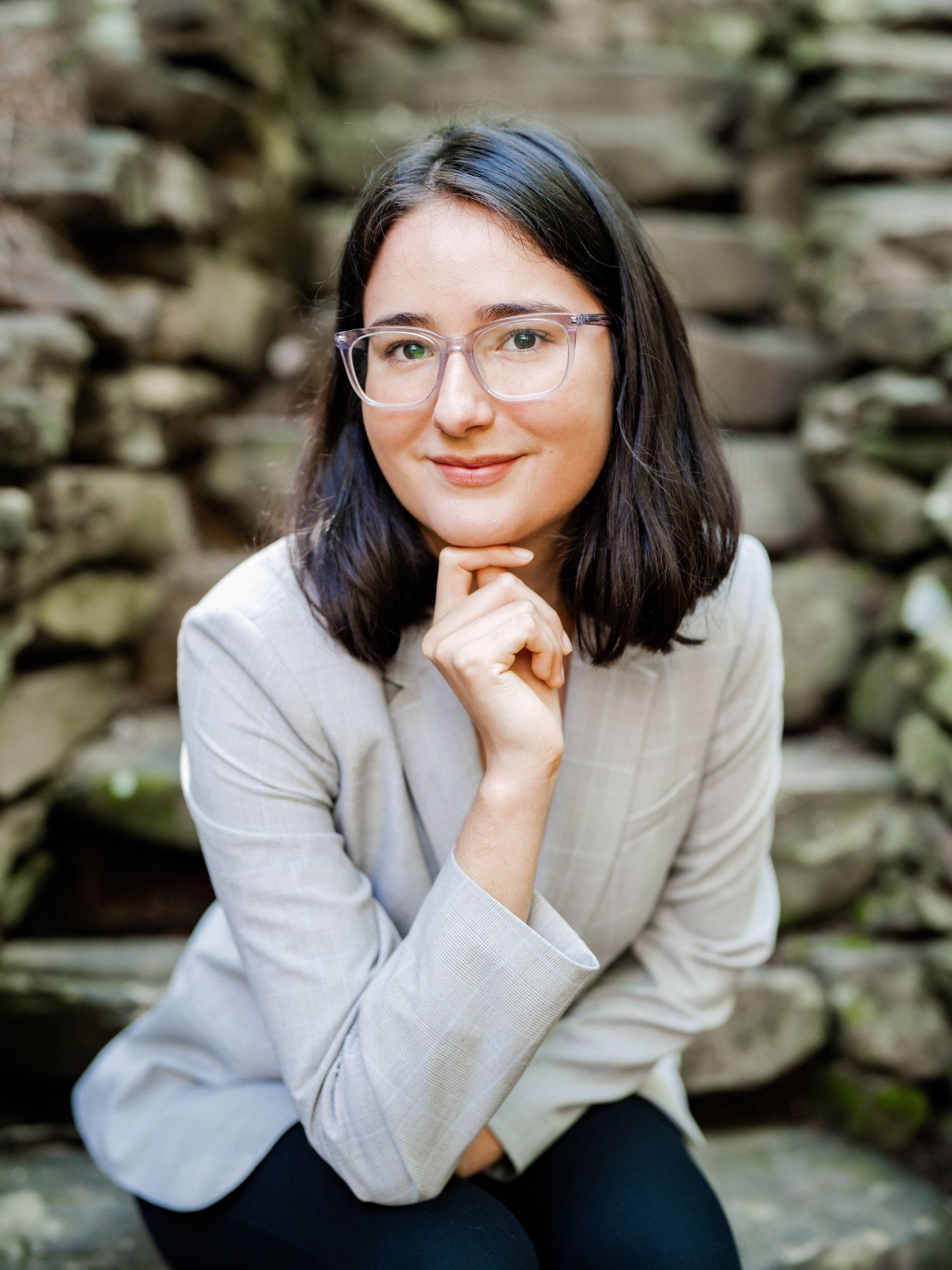 Professional portrait against a stone wall backdrop featuring someone in a light gray blazer and glasses with a contemplative pose.