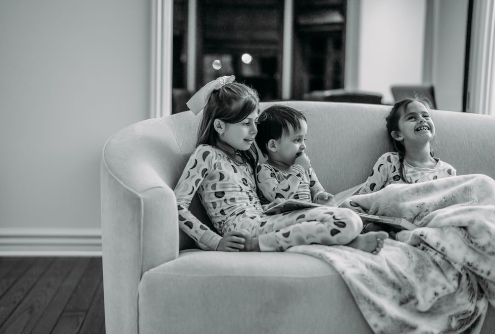 A black and white photograph showing three people relaxing together on a couch under blankets.
