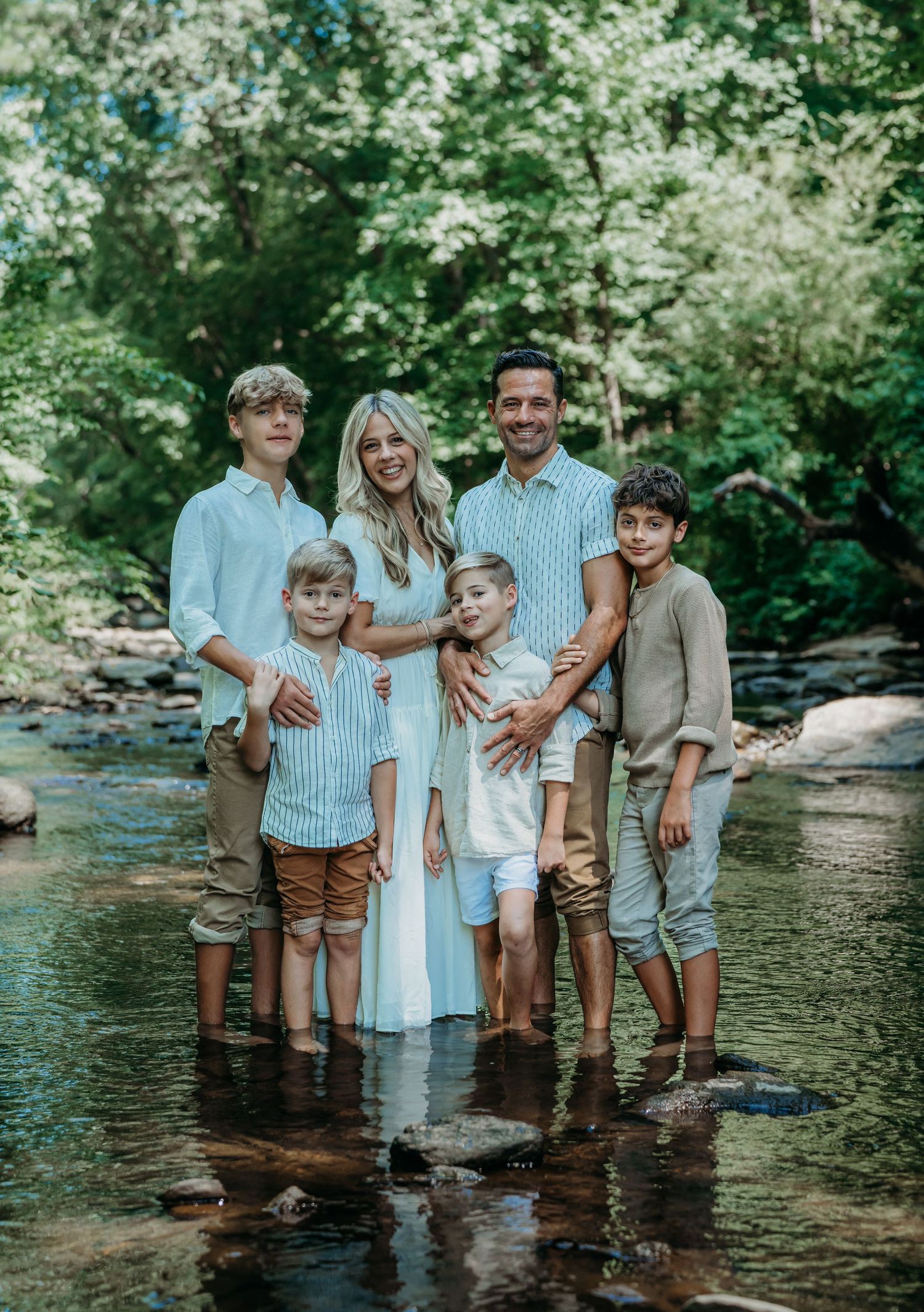 A family poses in matching blue and neutral tones while standing in a shallow creek surrounded by green trees.