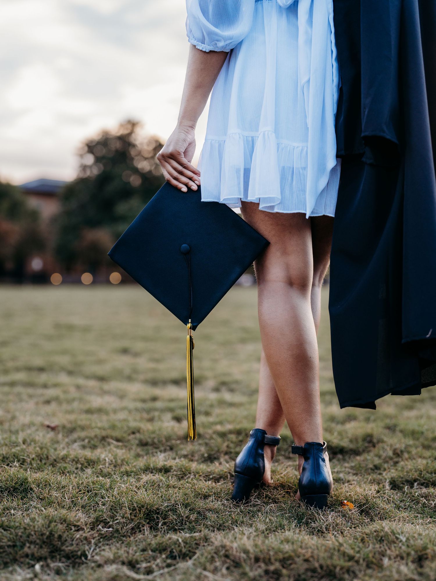 Graduate in light blue dress holds their graduation cap on green grass field while wearing navy blue shoes.