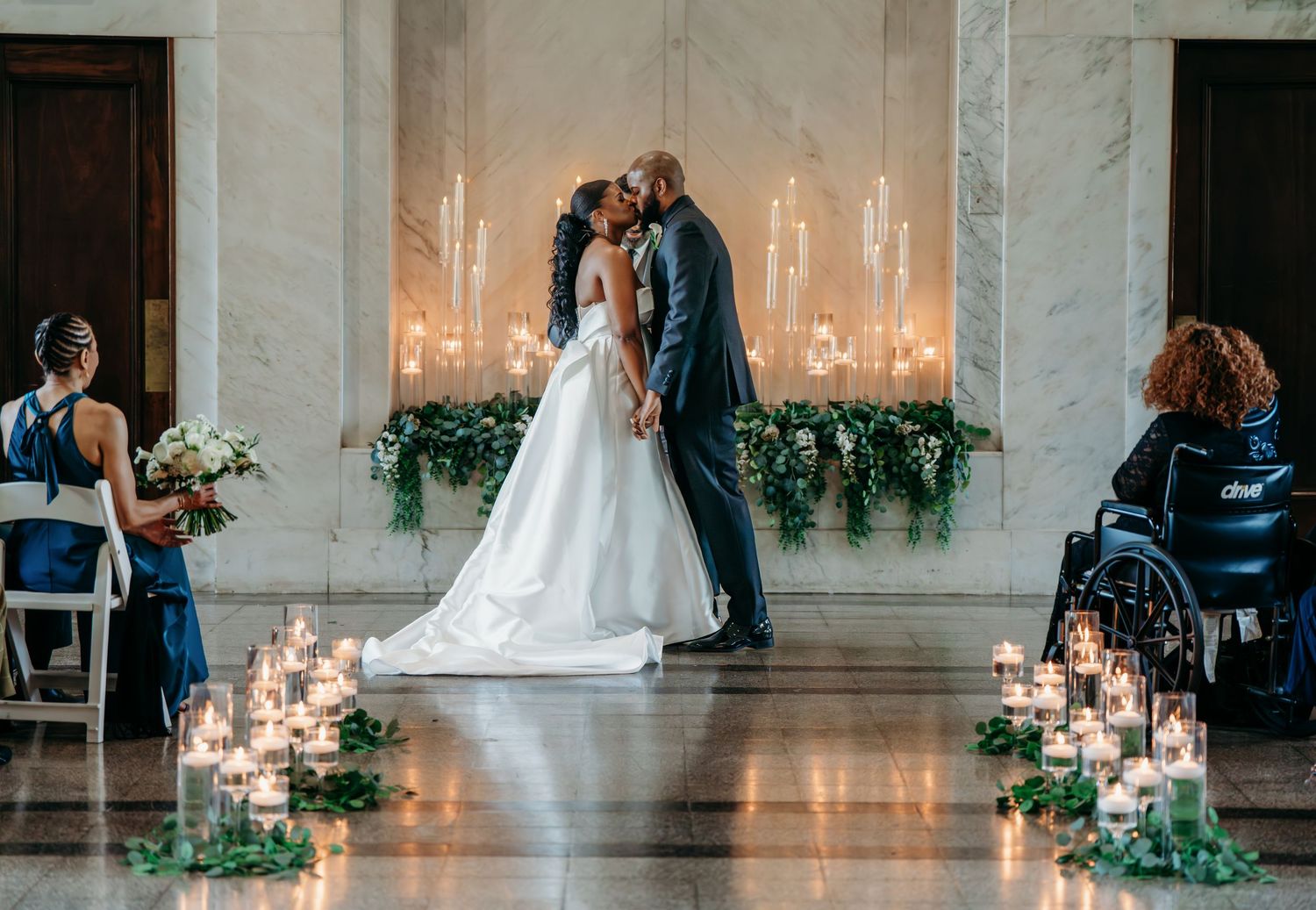 A romantic wedding ceremony in a modern venue with glowing candles and greenery lining the aisle and altar.