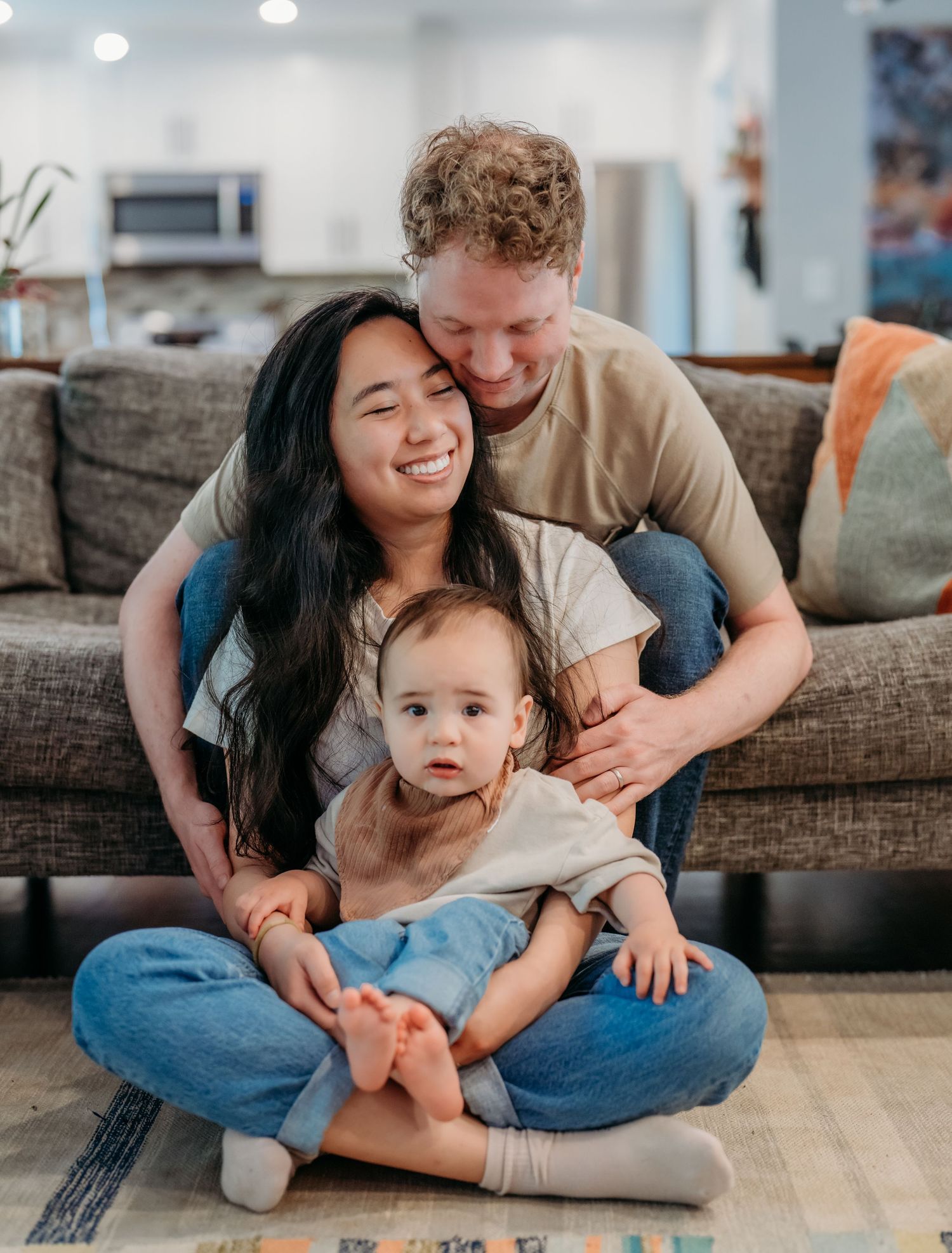 A happy family sits together on a gray couch in their living room, sharing joyful laughter and embraces.