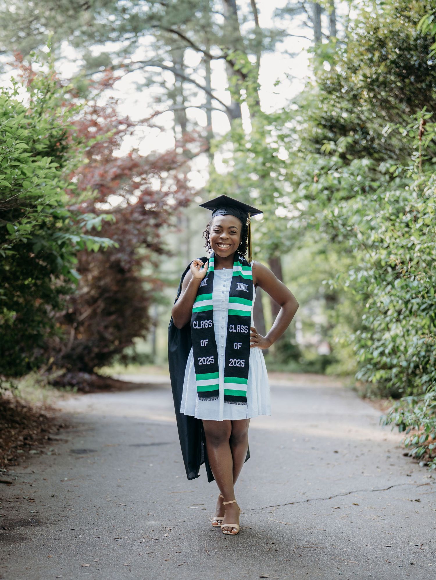 Graduation portrait of a person in cap and gown with traditional stole on a tree-lined pathway.