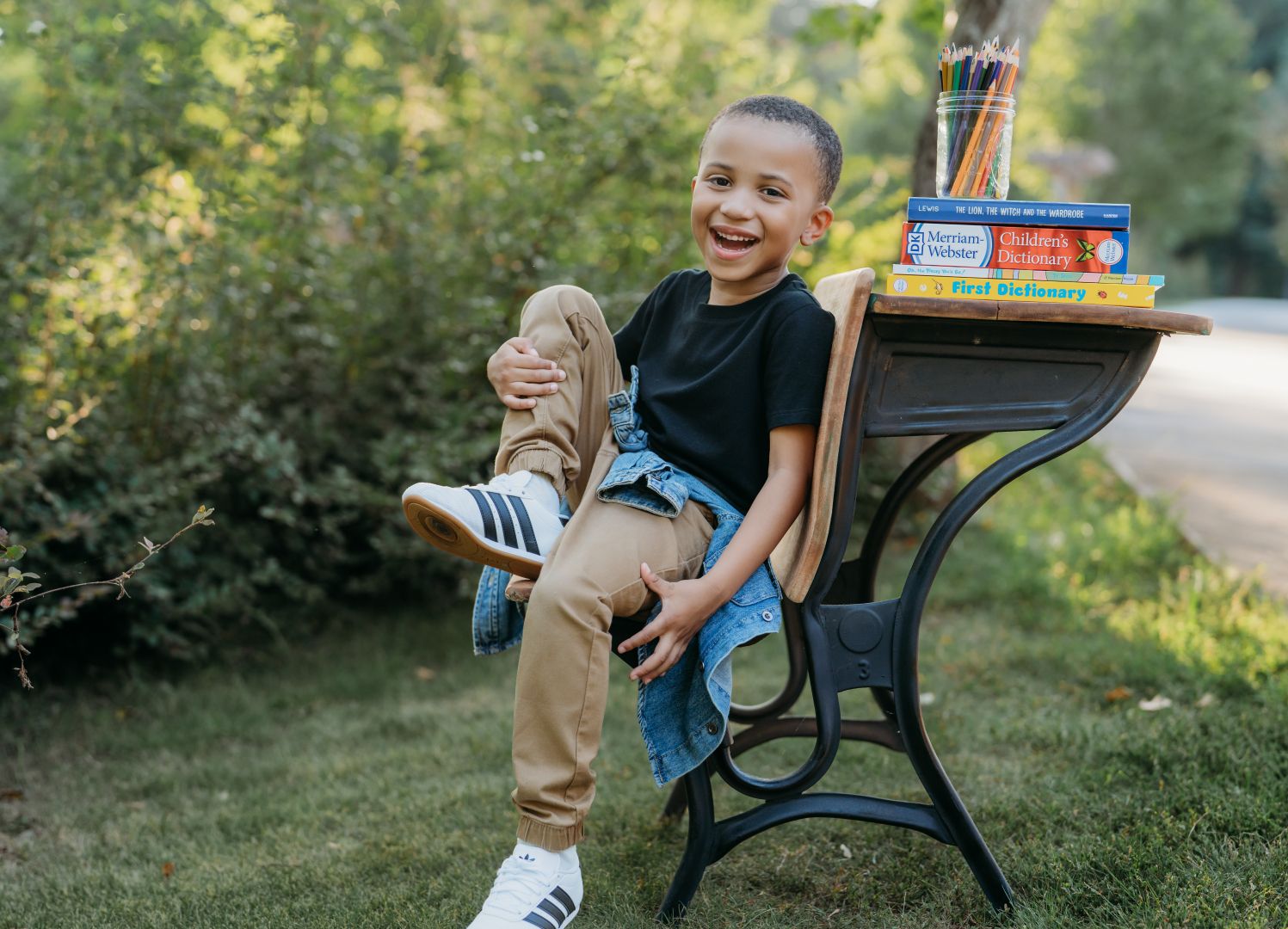 A smiling young boy sits on a school desk outdoors, wearing a black shirt and khaki pants with white sneakers.