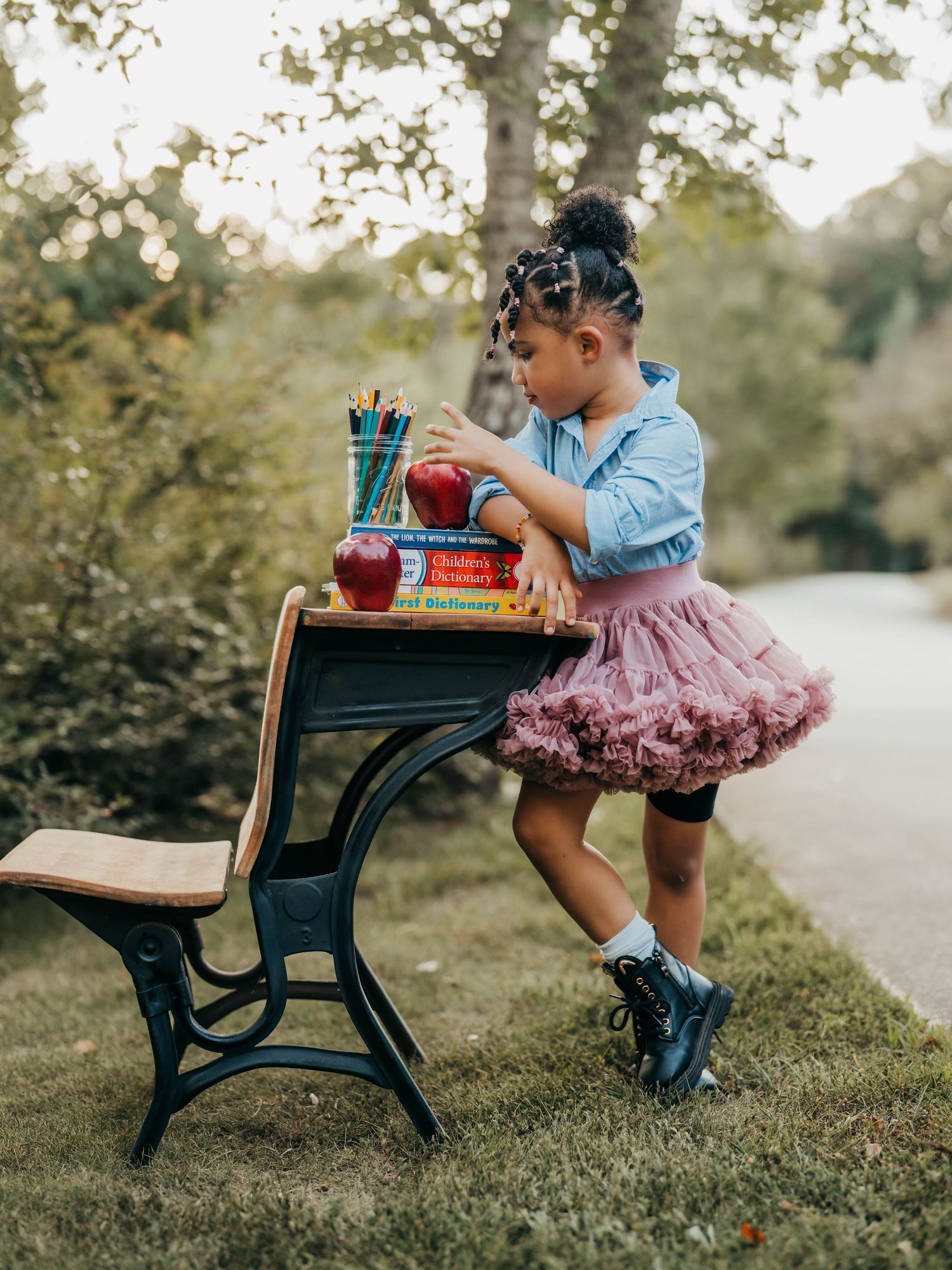 A young girl in a pink tutu and denim shirt sits at a vintage school desk outdoors with books and pencils.