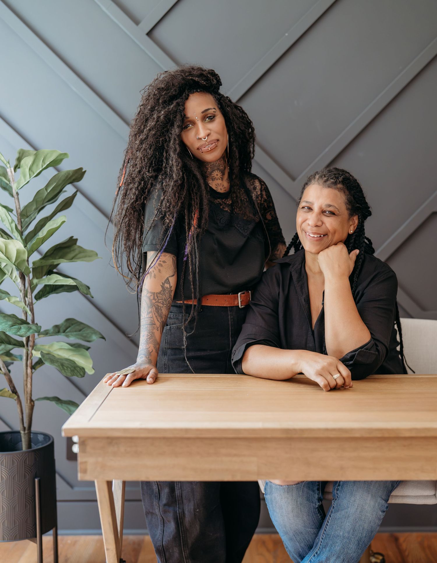 Two women pose confidently at a wooden table against a geometric gray wall, one standing with tattoos visible.