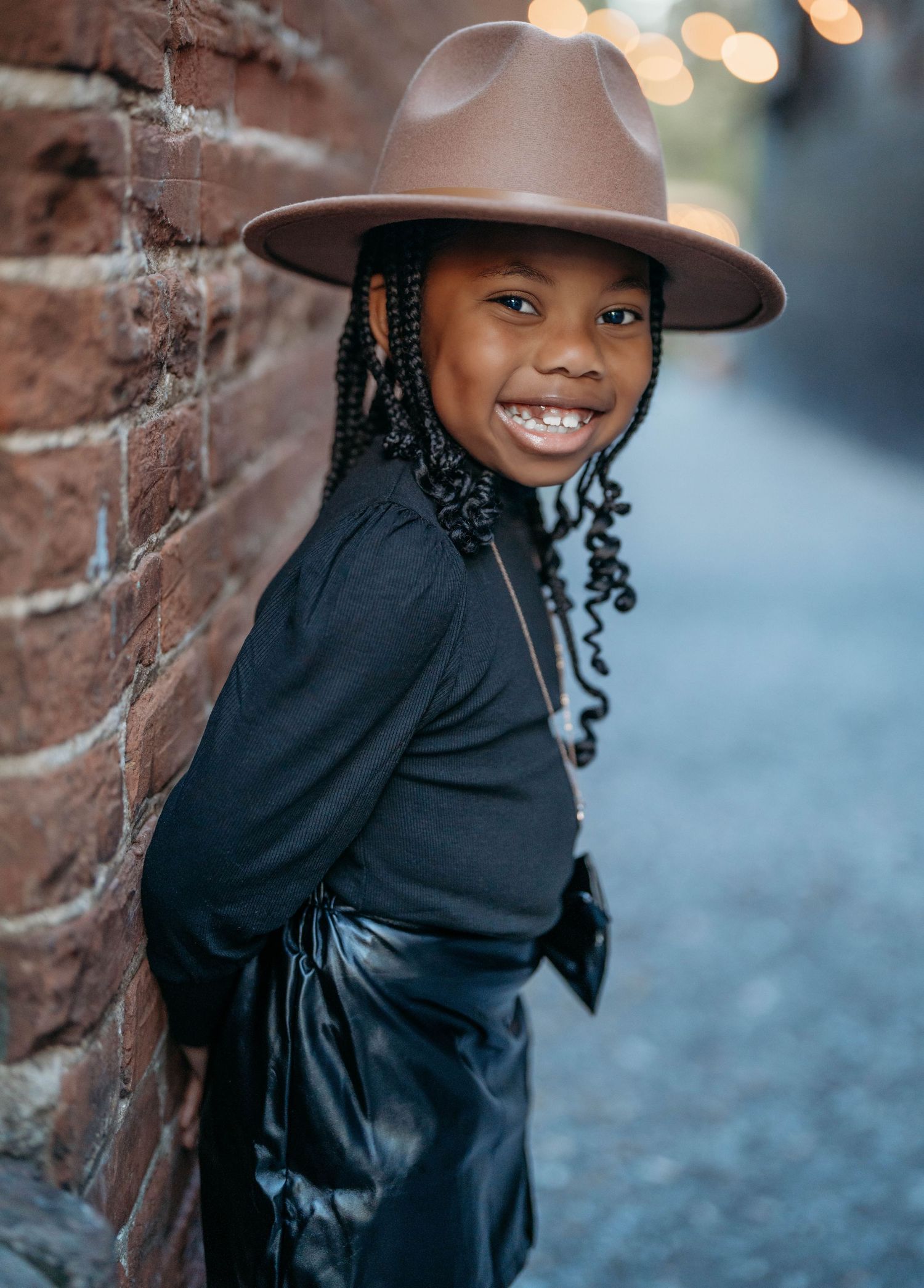 A young girl in a black outfit poses playfully against a brick wall with bokeh lights in the background.
