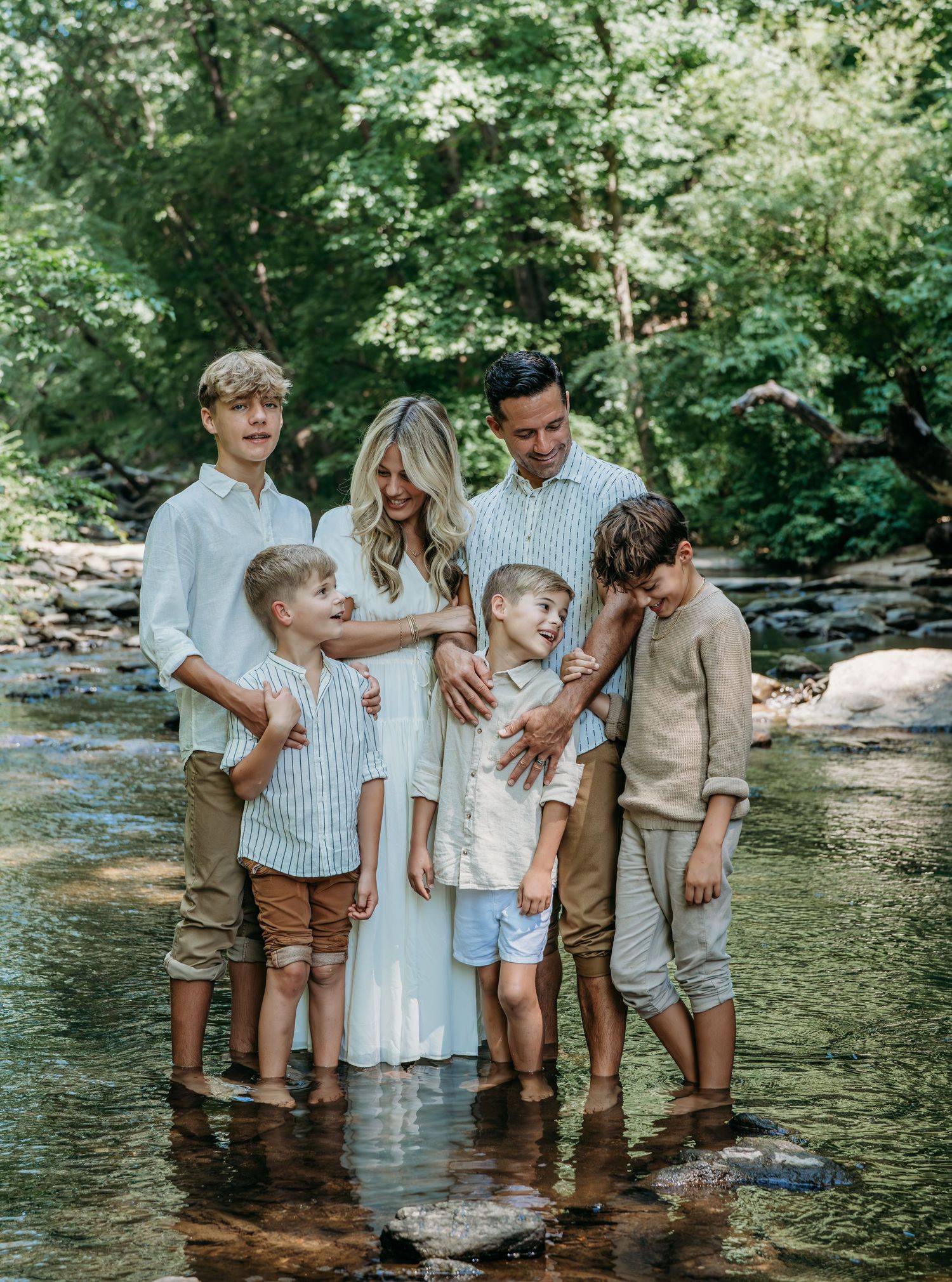 Family in coordinated neutral outfits stands together in a shallow creek surrounded by lush green forest scenery.