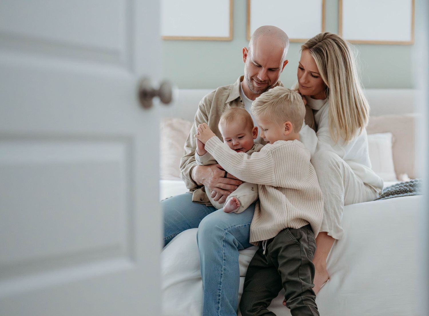 A candid family moment showing parents and baby interacting in a bright, modern bedroom setting. Shot through an open door