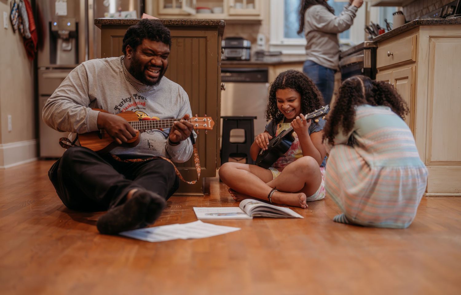 A family sits on a hardwood floor playing ukuleles together in a cozy kitchen setting.