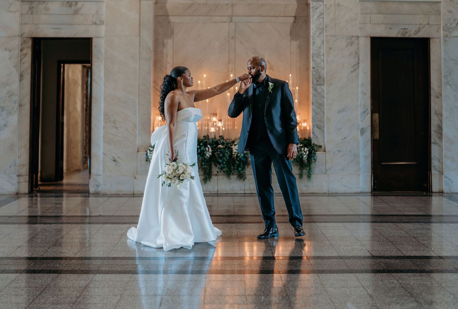 A couple in formal wedding attire dances together in an elegant marble hallway with warm lighting.