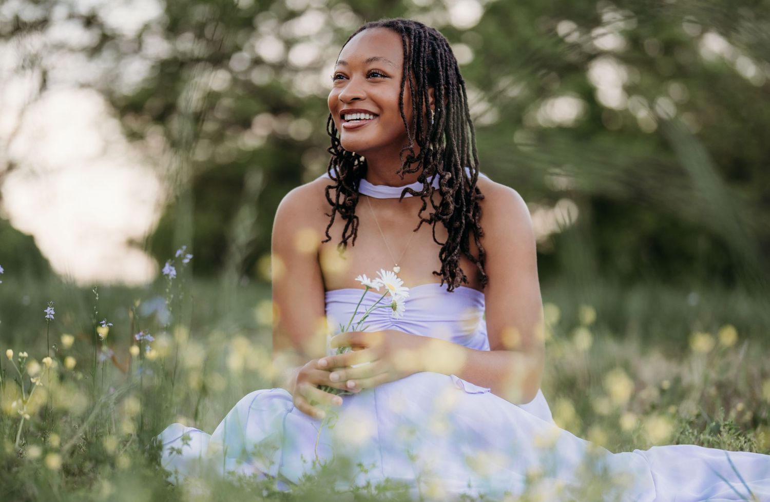 A smiling young woman with braids sits in a wildflower meadow holding a daisy in a light purple dress.