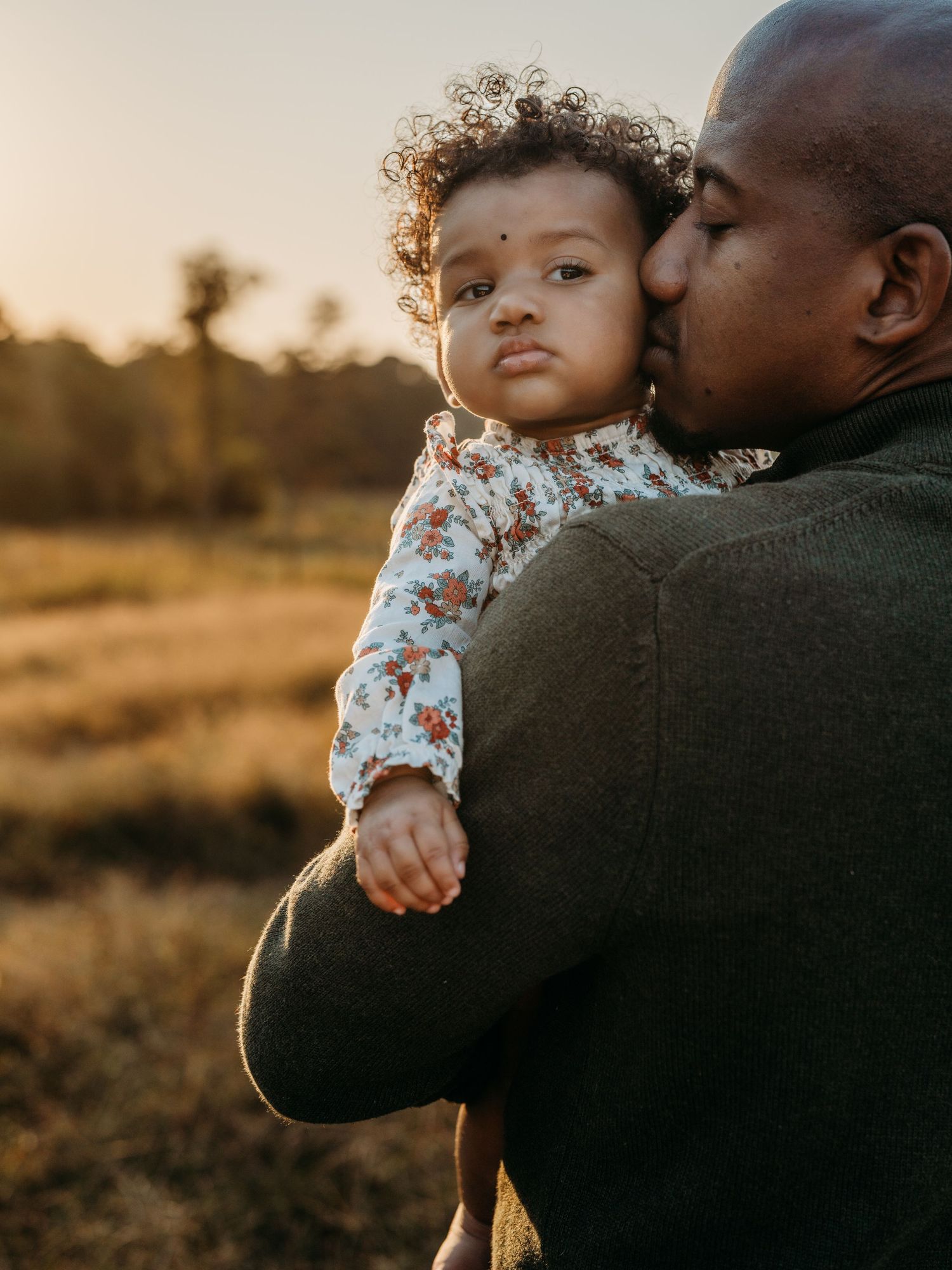 Tender moment captured at sunset as a parent in a dark sweater embraces and kisses their curly-haired child in floral print.