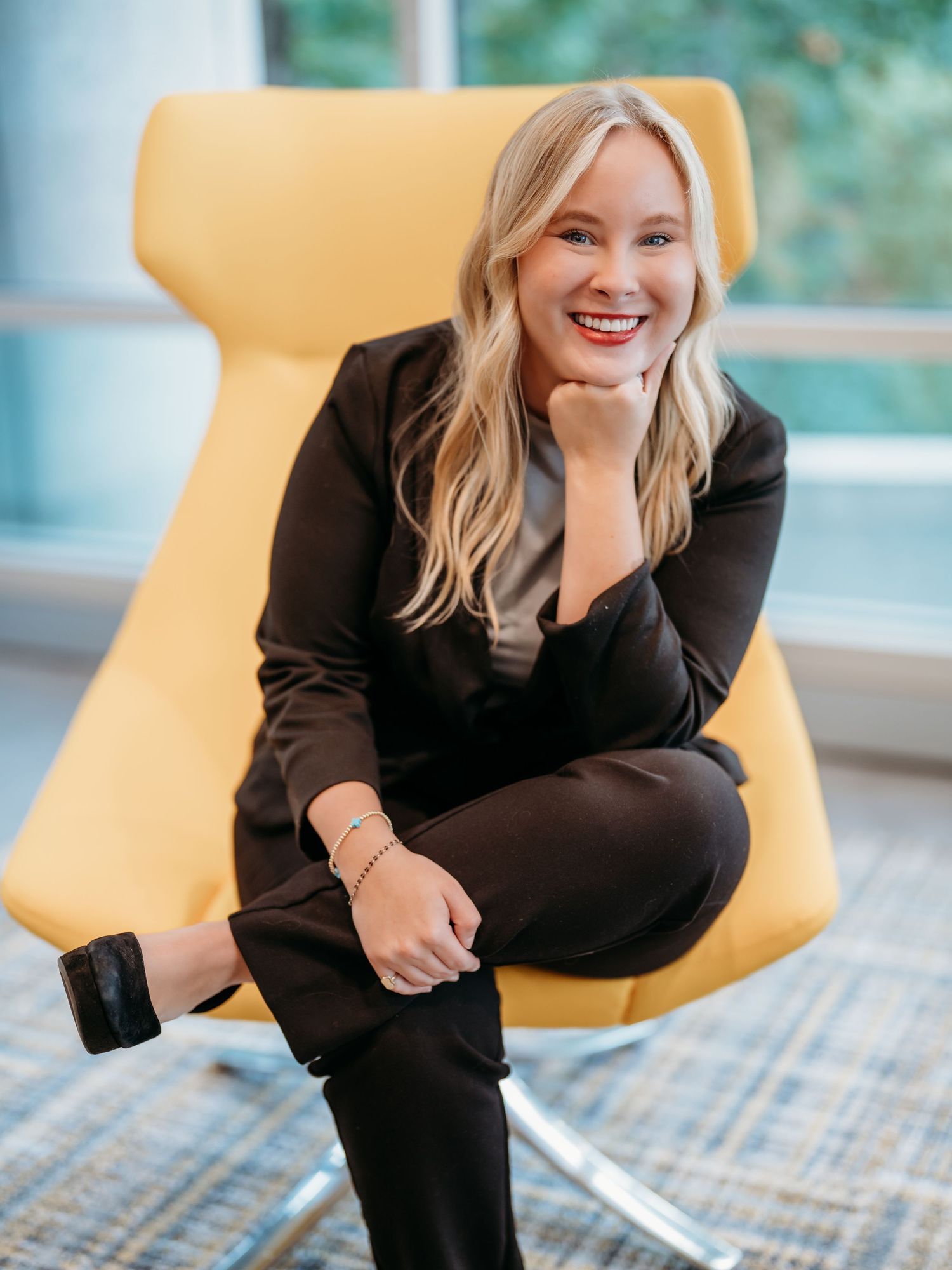 Professional wearing black blazer sitting in yellow accent chair displays joyful expression during business photoshoot.