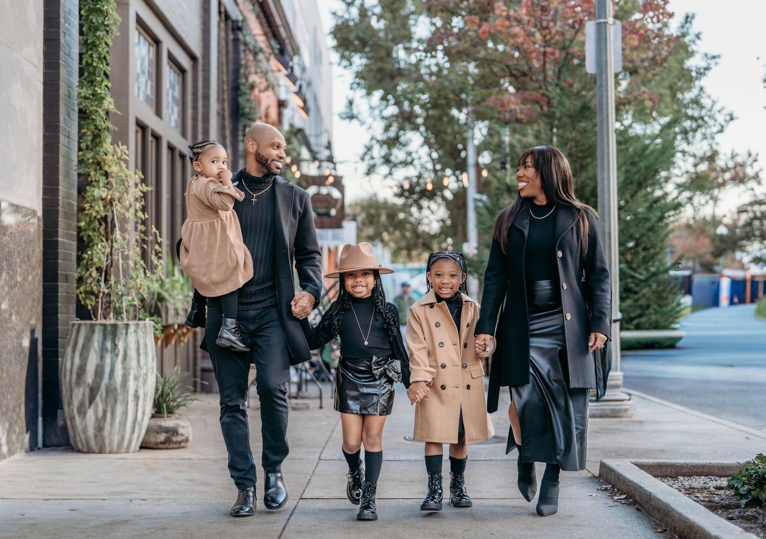 A stylish family walks together on an urban sidewalk wearing coordinated fall outfits on a sunny autumn day.