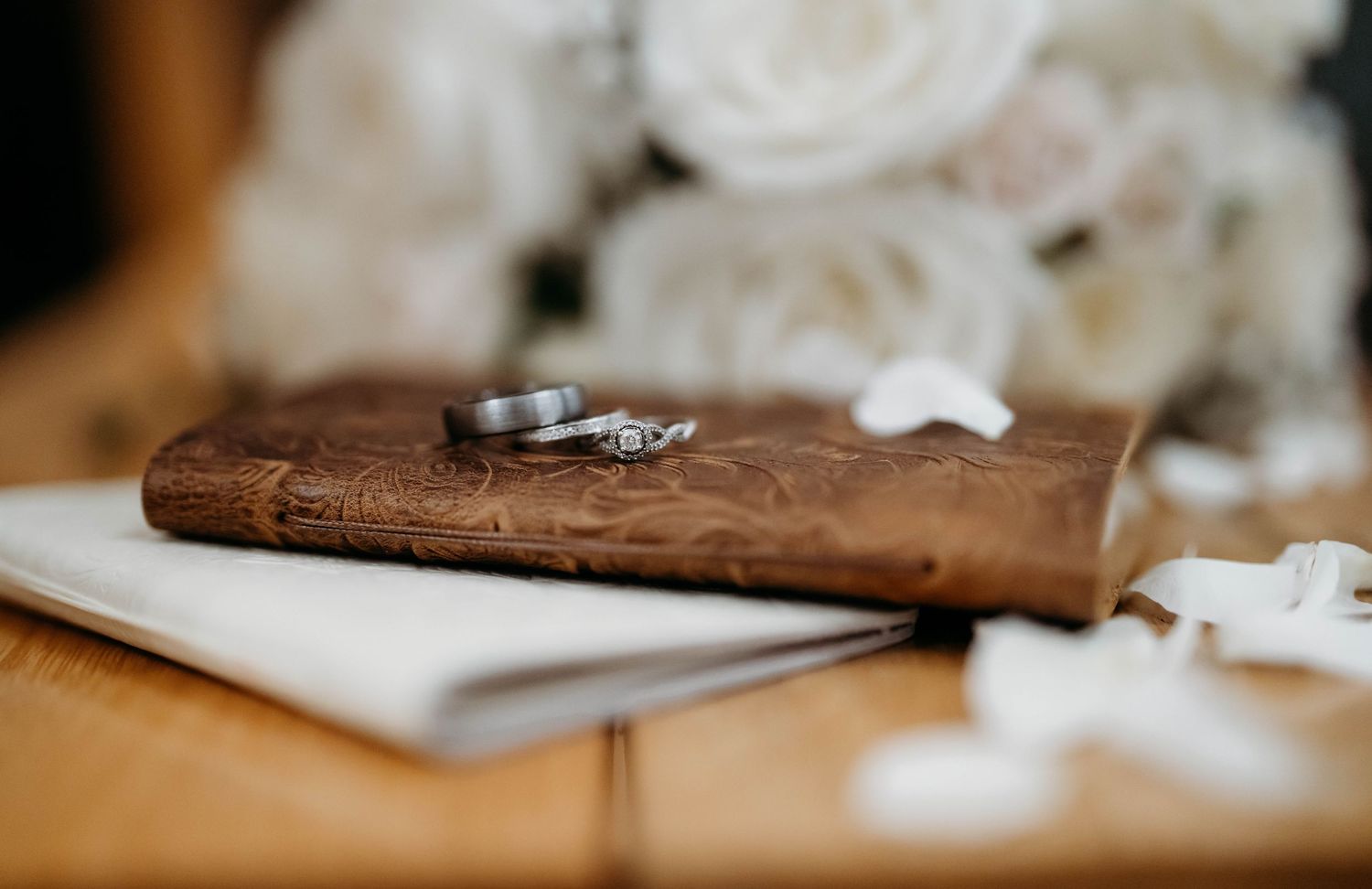 A picture of leather vow book, flowers in the background and wedding rings sitting on top of the book