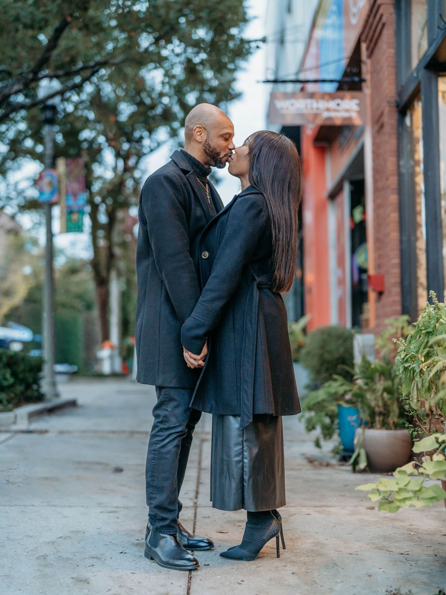 A couple shares a romantic kiss while standing on a brick-lined sidewalk near potted plants in an urban setting.