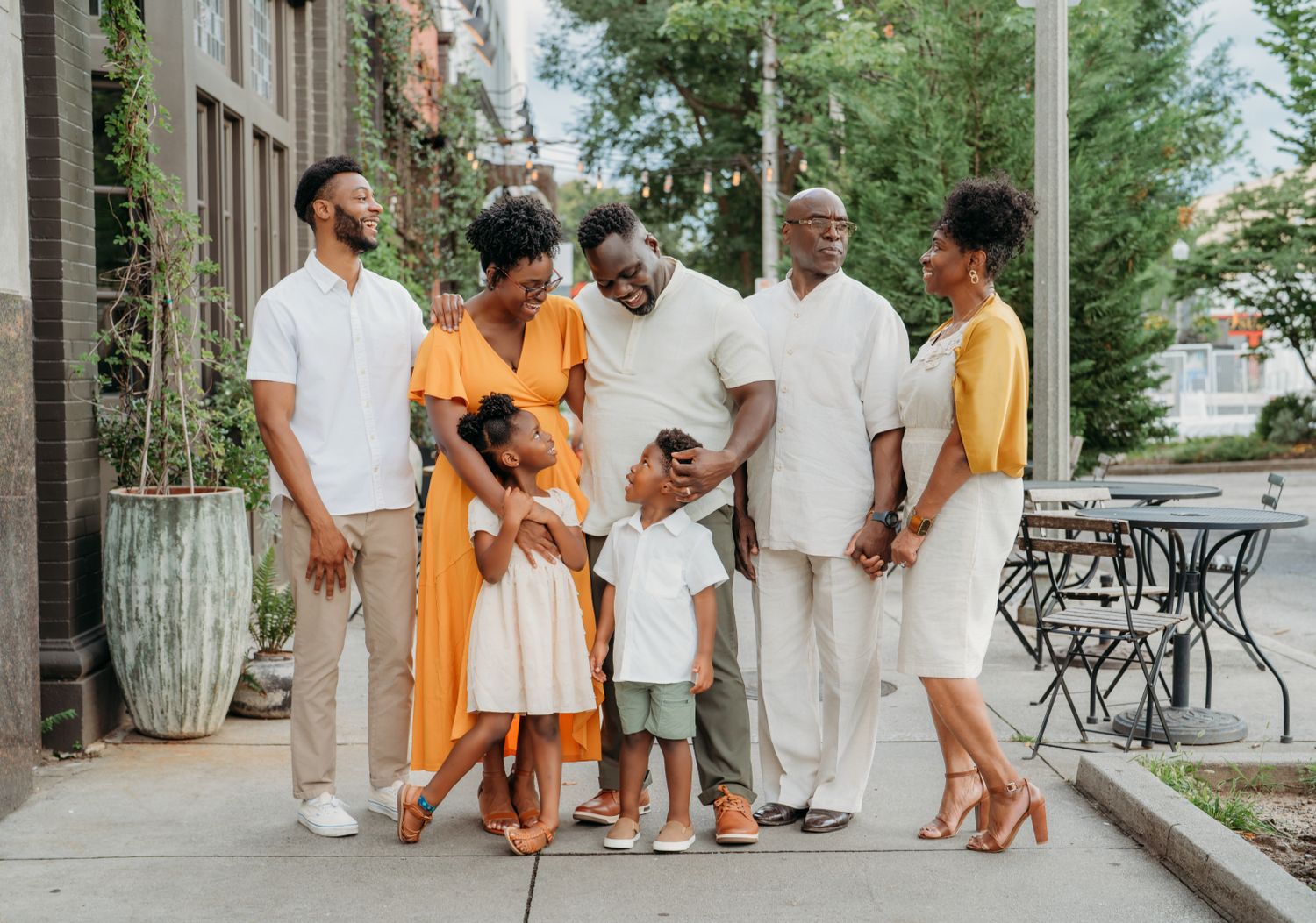 A joyful multigenerational Black family poses together outdoors on a sunny sidewalk, dressed in elegant white and orange outfits.