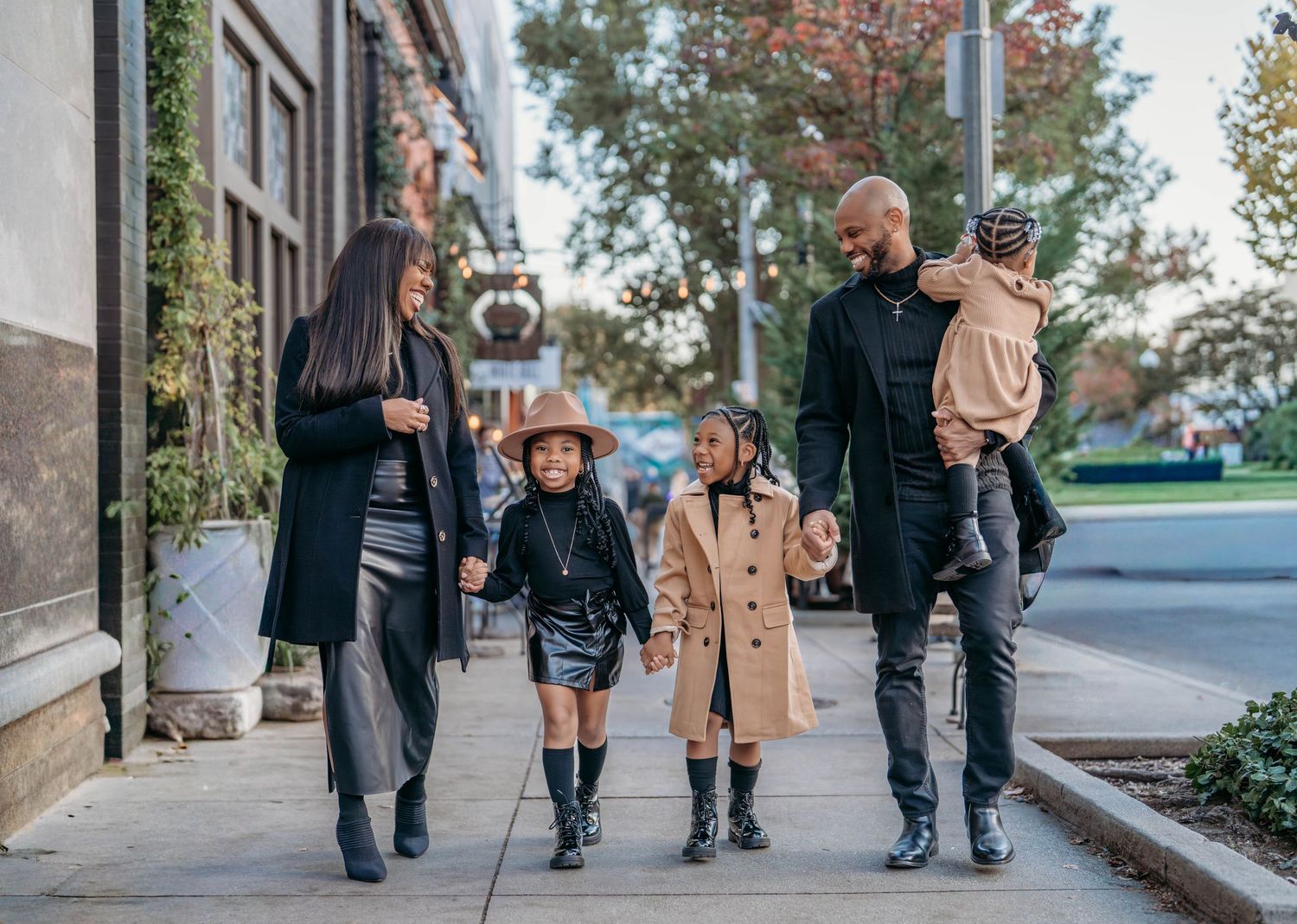 A young family walking together on a city sidewalk during autumn with trees and buildings in the background.