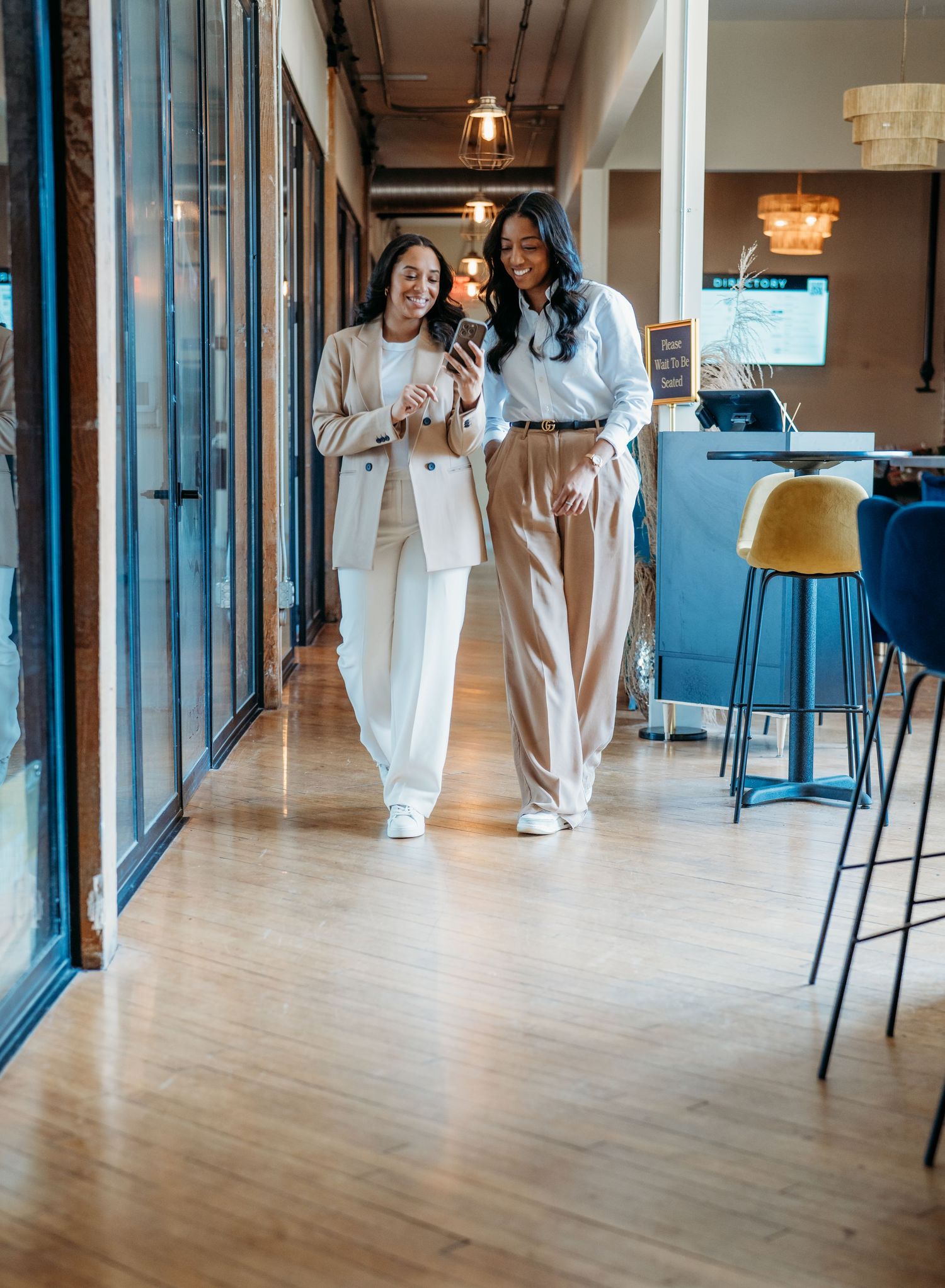 Two coworkers in coordinated neutral outfits look at a mobile phone while walking in a modern office space.