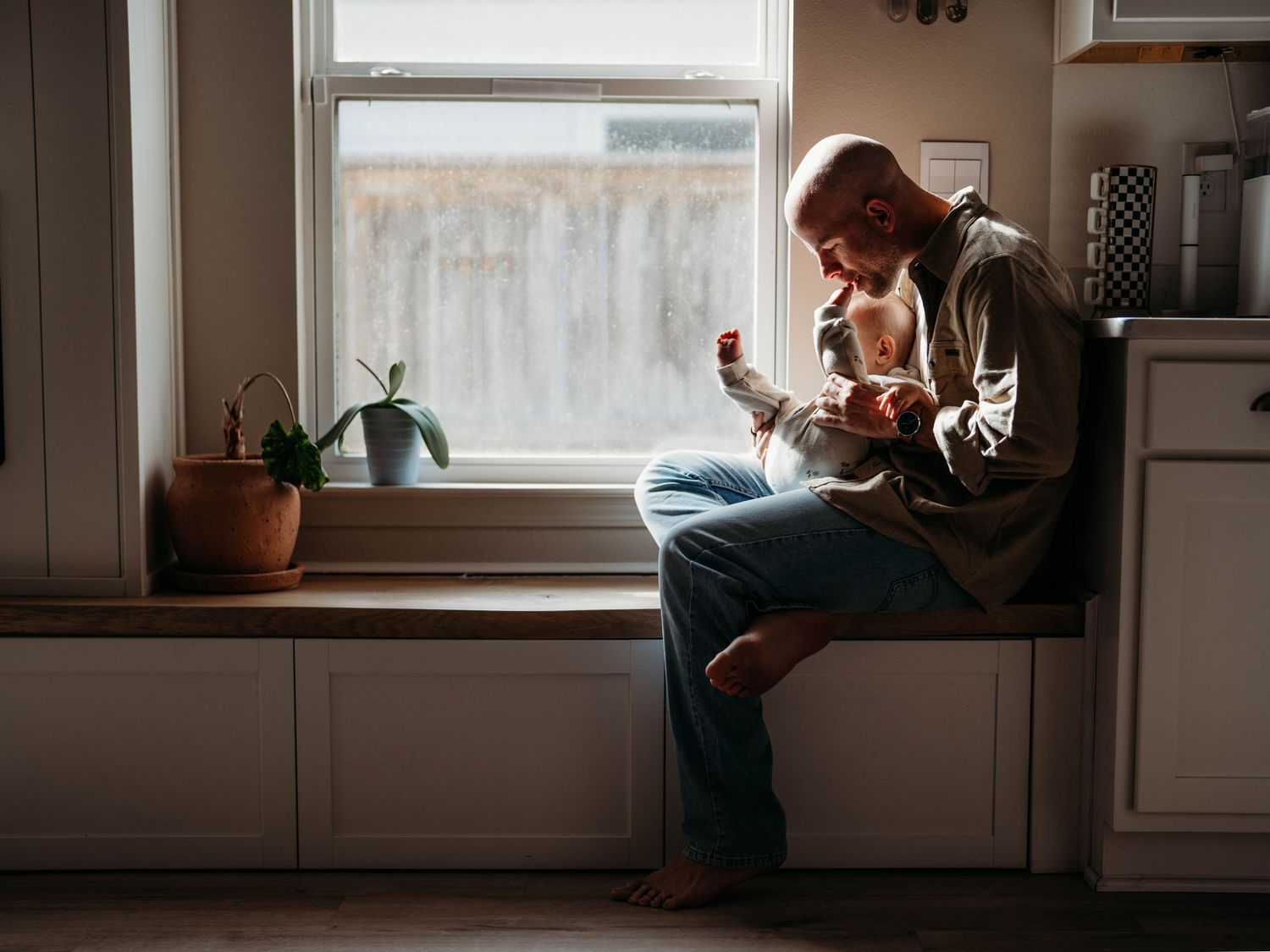 A photos showing a father sitting by a window while playing with his baby in a cozy interior setting.
