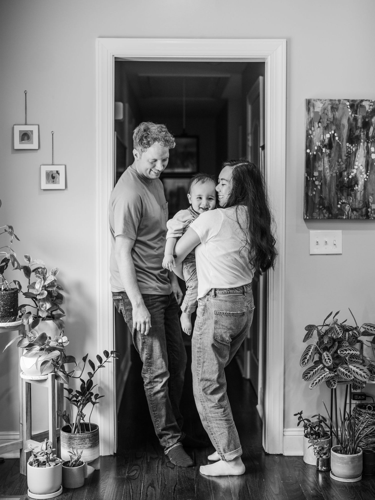 A couple stands in a doorway with a small baby, surrounded by houseplants in a monochrome domestic scene.